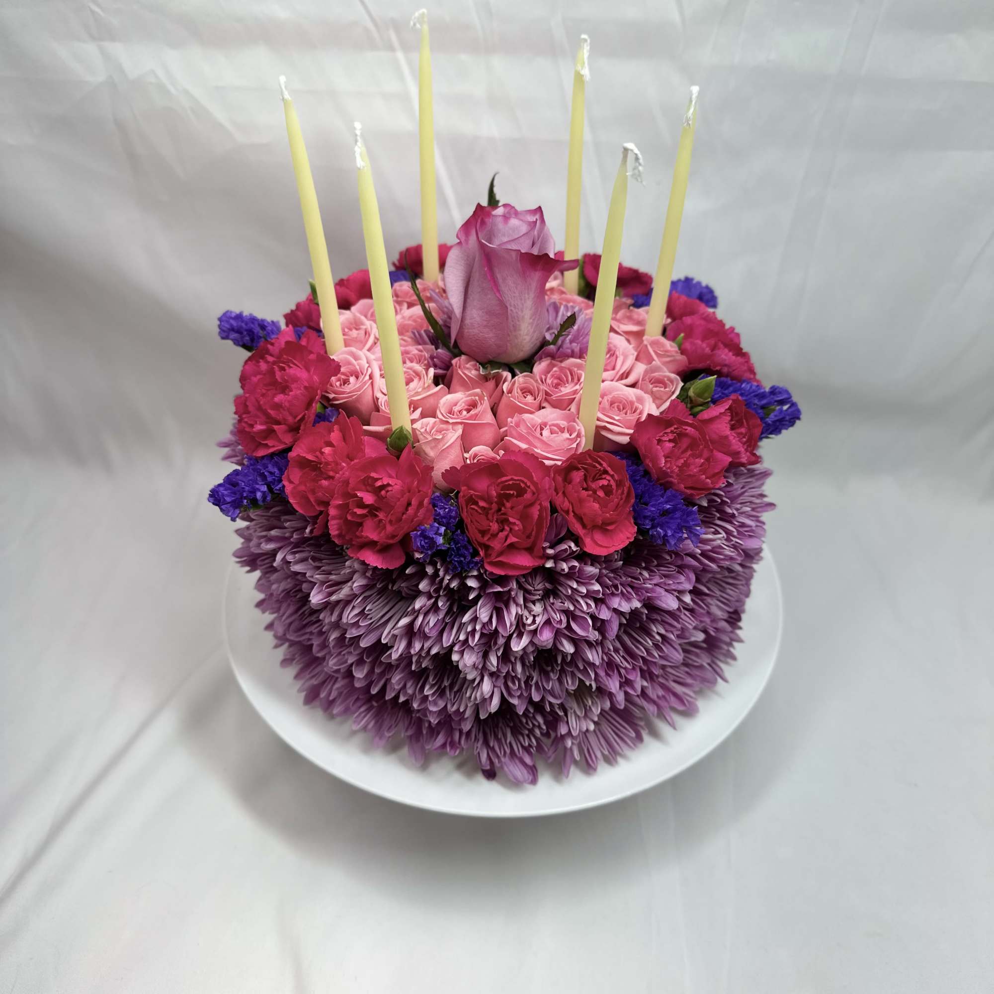 Round floral cake with pink roses, purple blooms, and tall candles on a white plate