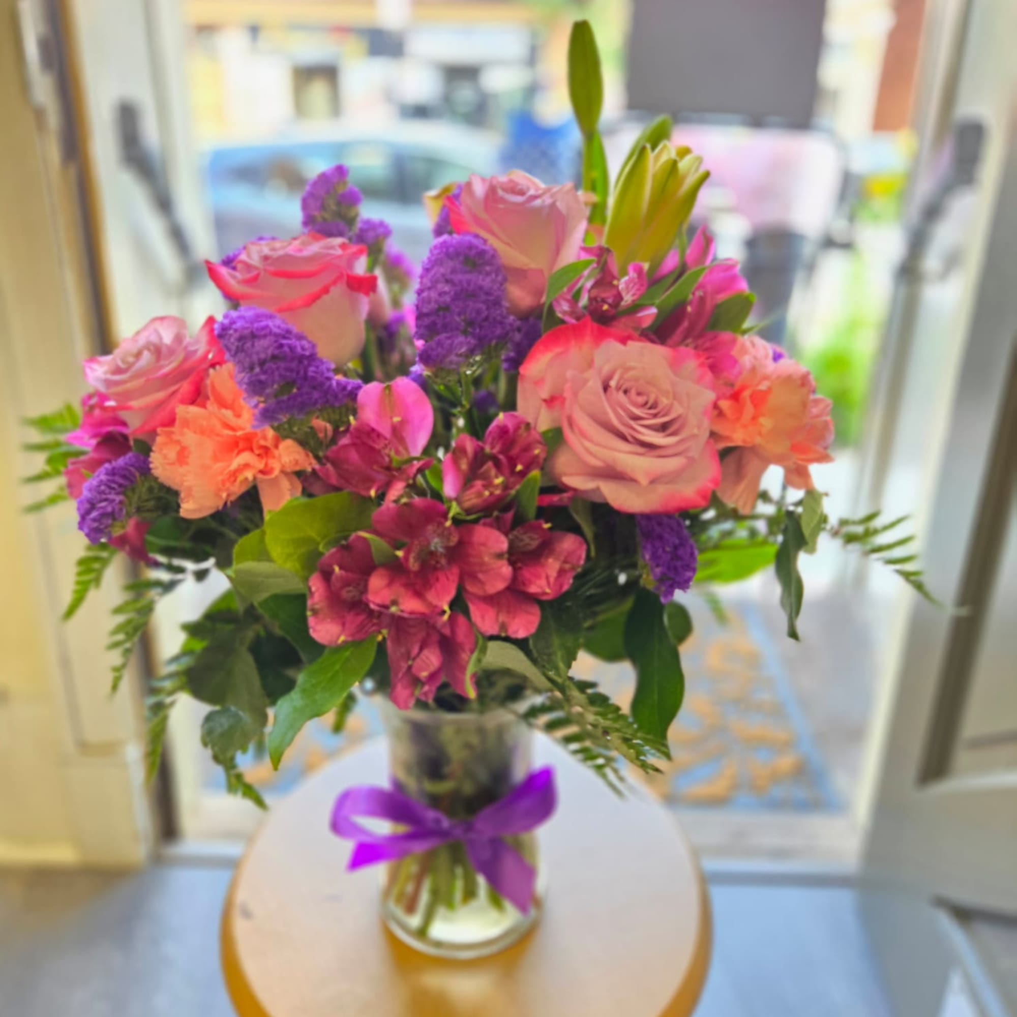 Bouquet of pink roses, purple flowers, and lilies in a glass vase with a purple ribbon
