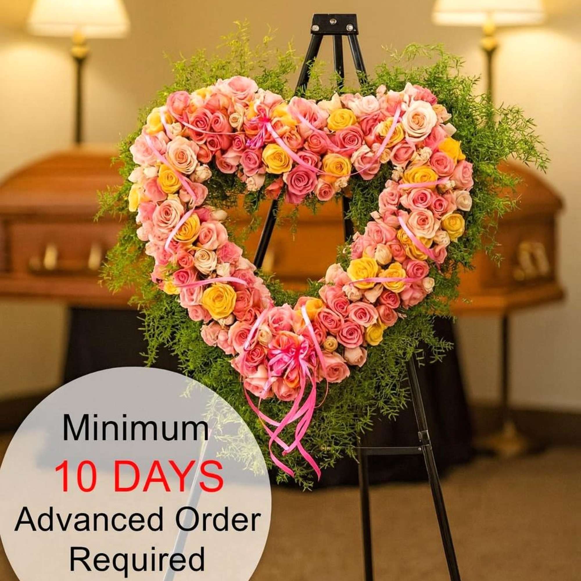 Heart-shaped wreath of pink and yellow roses on a metal easel in a funeral setting.