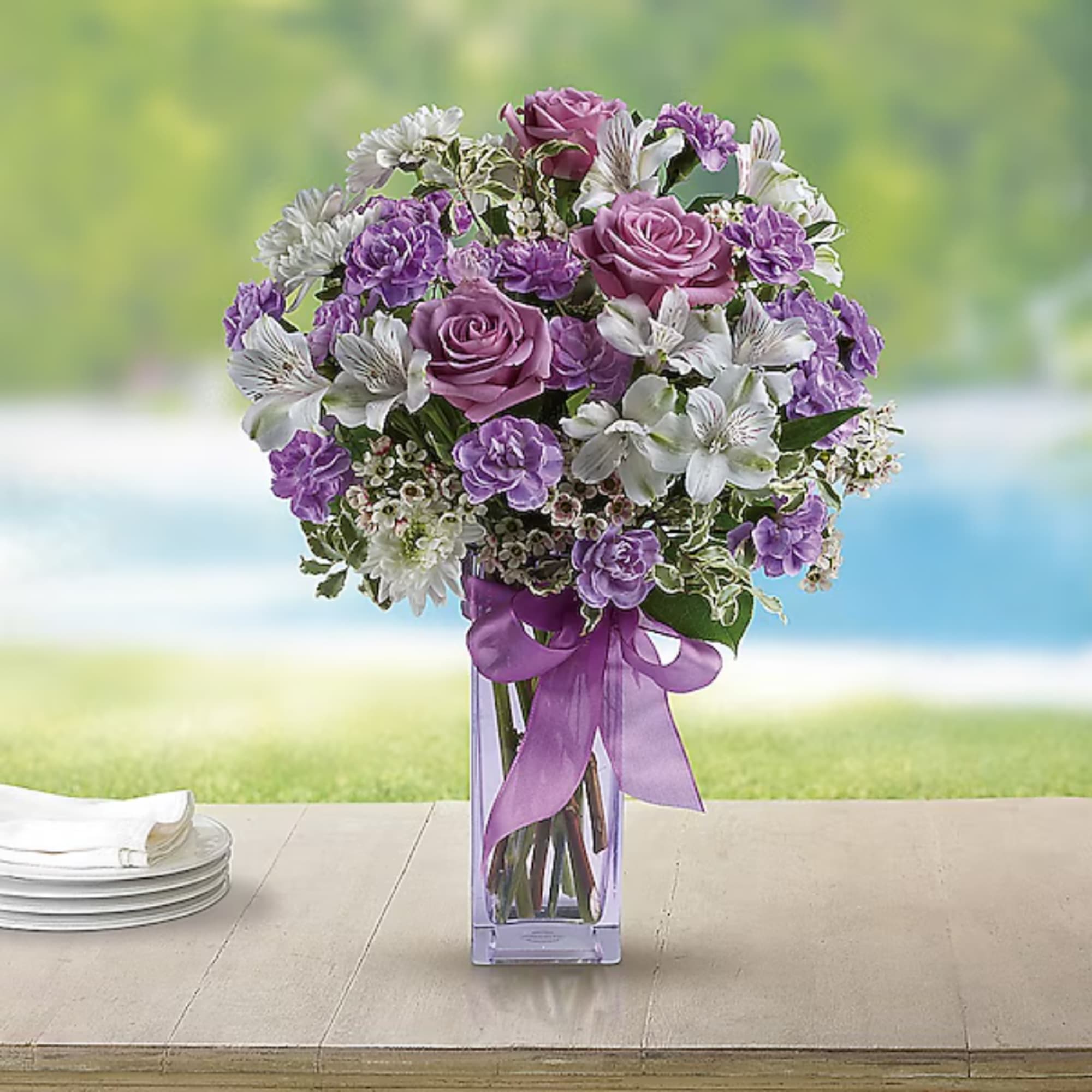 Purple and white bouquet in a clear square vase with a ribbon
