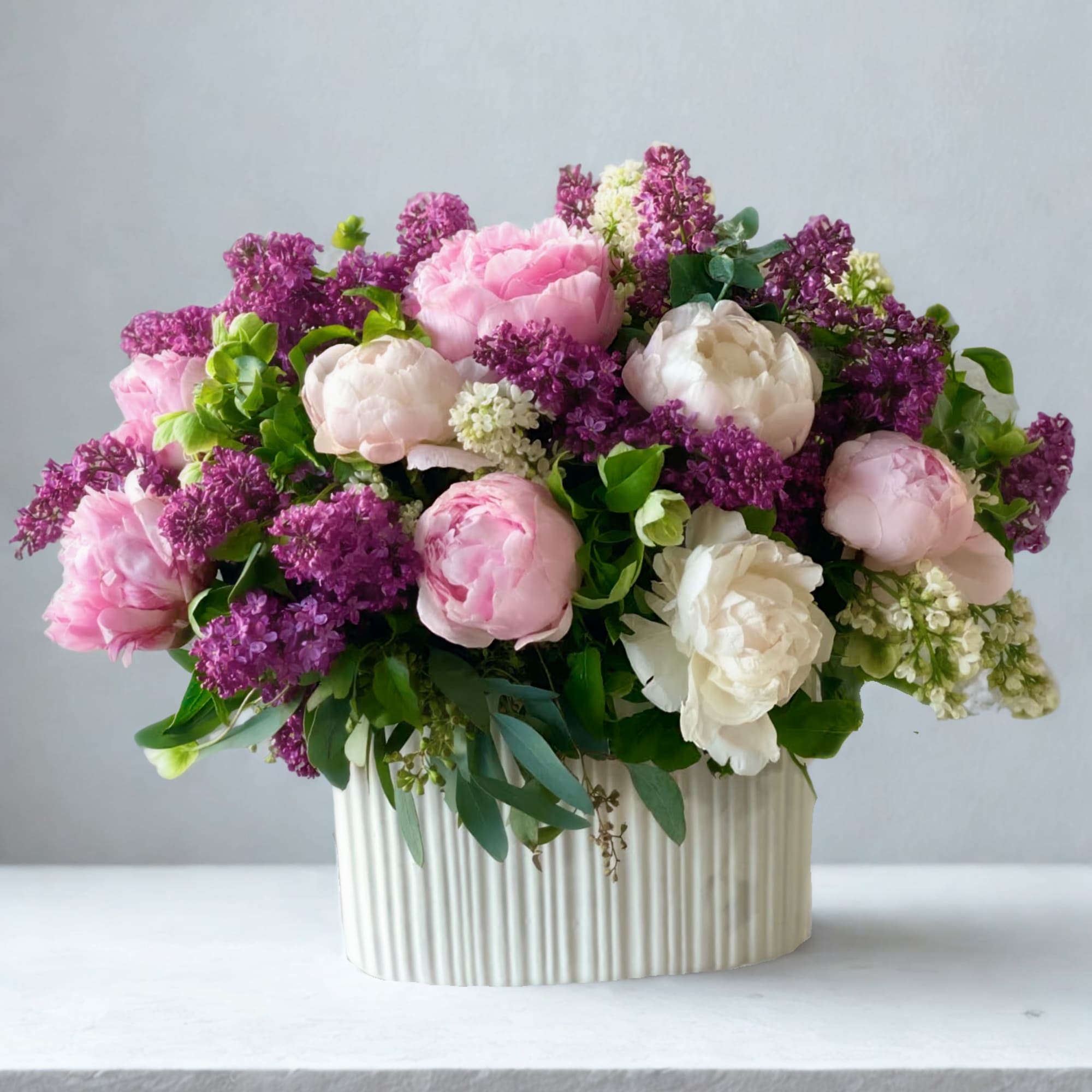 Pink and white peonies with purple lilac in a ribbed cream vase