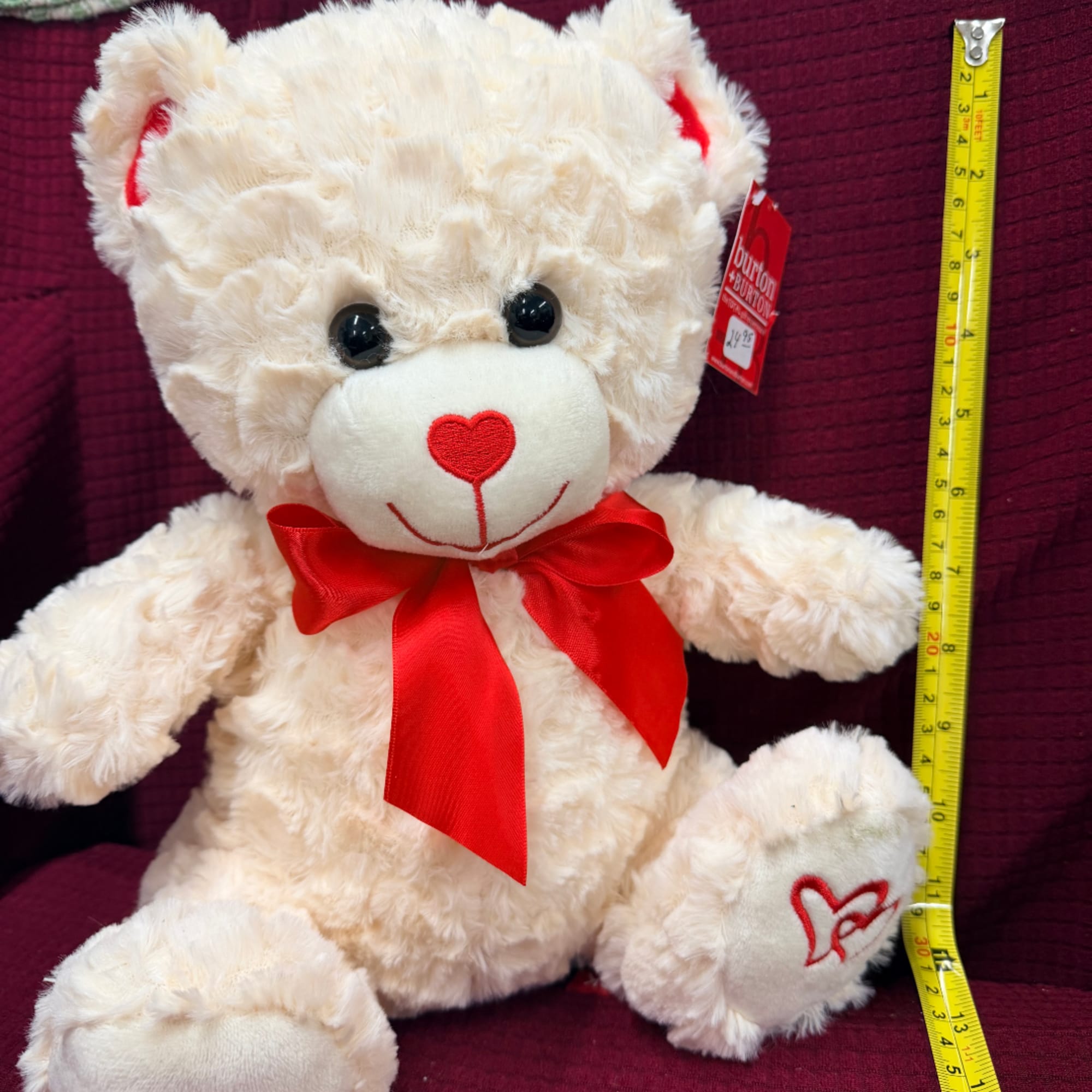 Cream teddy bear with a red bow and heart embroidery, shown with a measuring tape