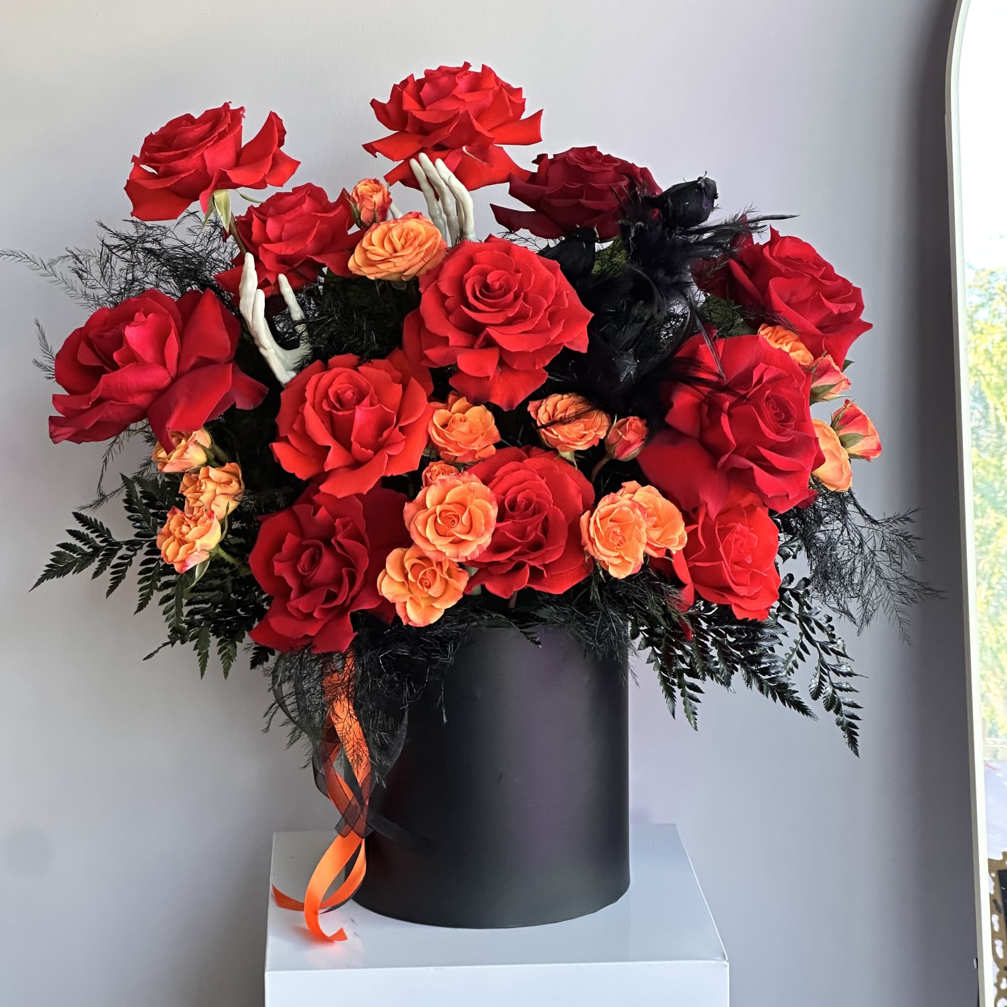 Dramatic arrangement of red and orange roses in a black cylinder with skeleton hands and dark accents.