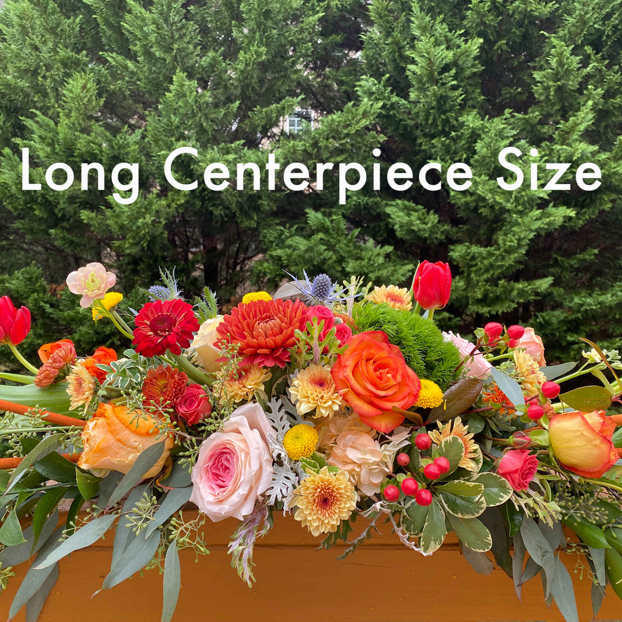 Long low centerpiece of mixed orange, red, yellow, and pink flowers in a wooden container.