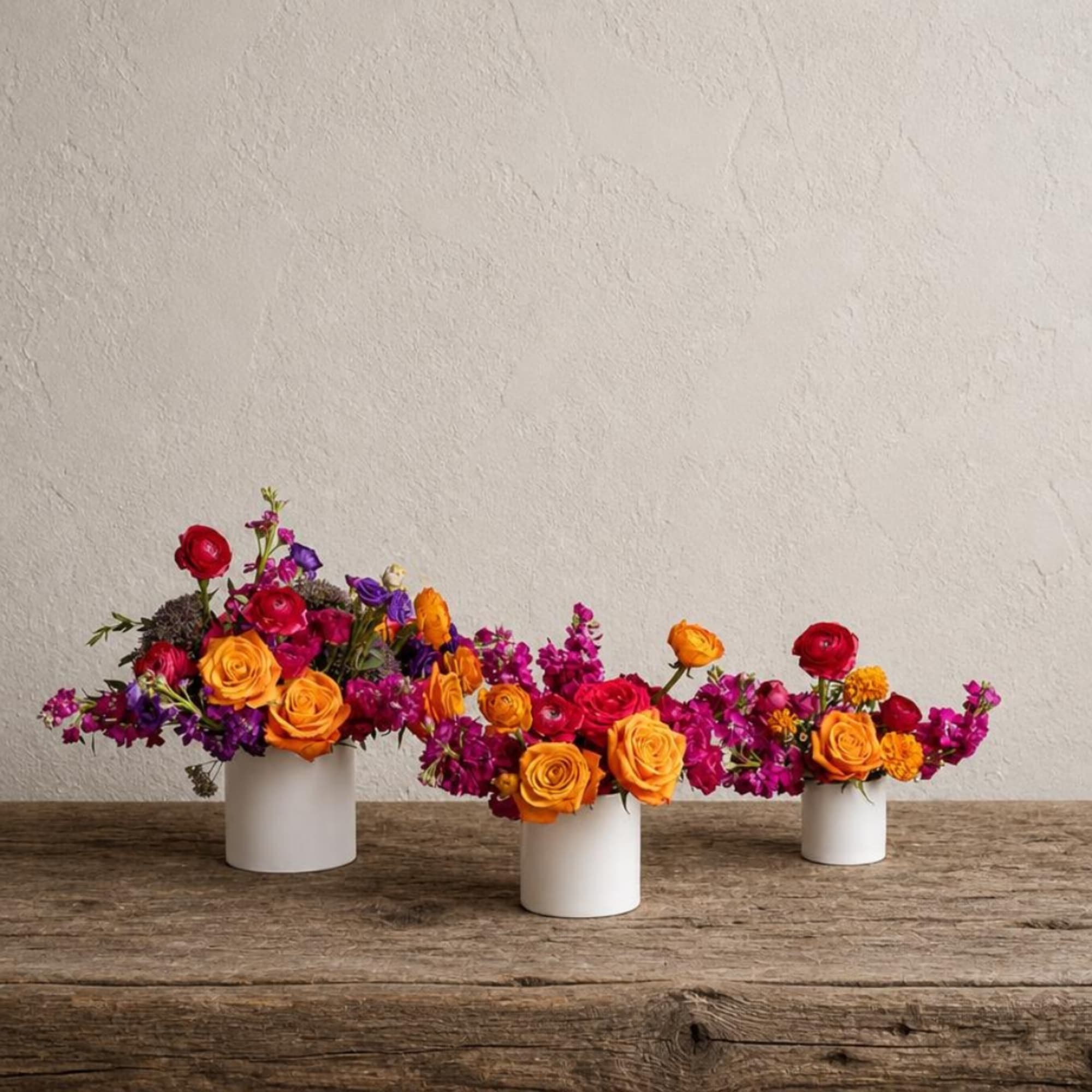 Three low floral arrangements in white containers with orange, red, and purple blooms