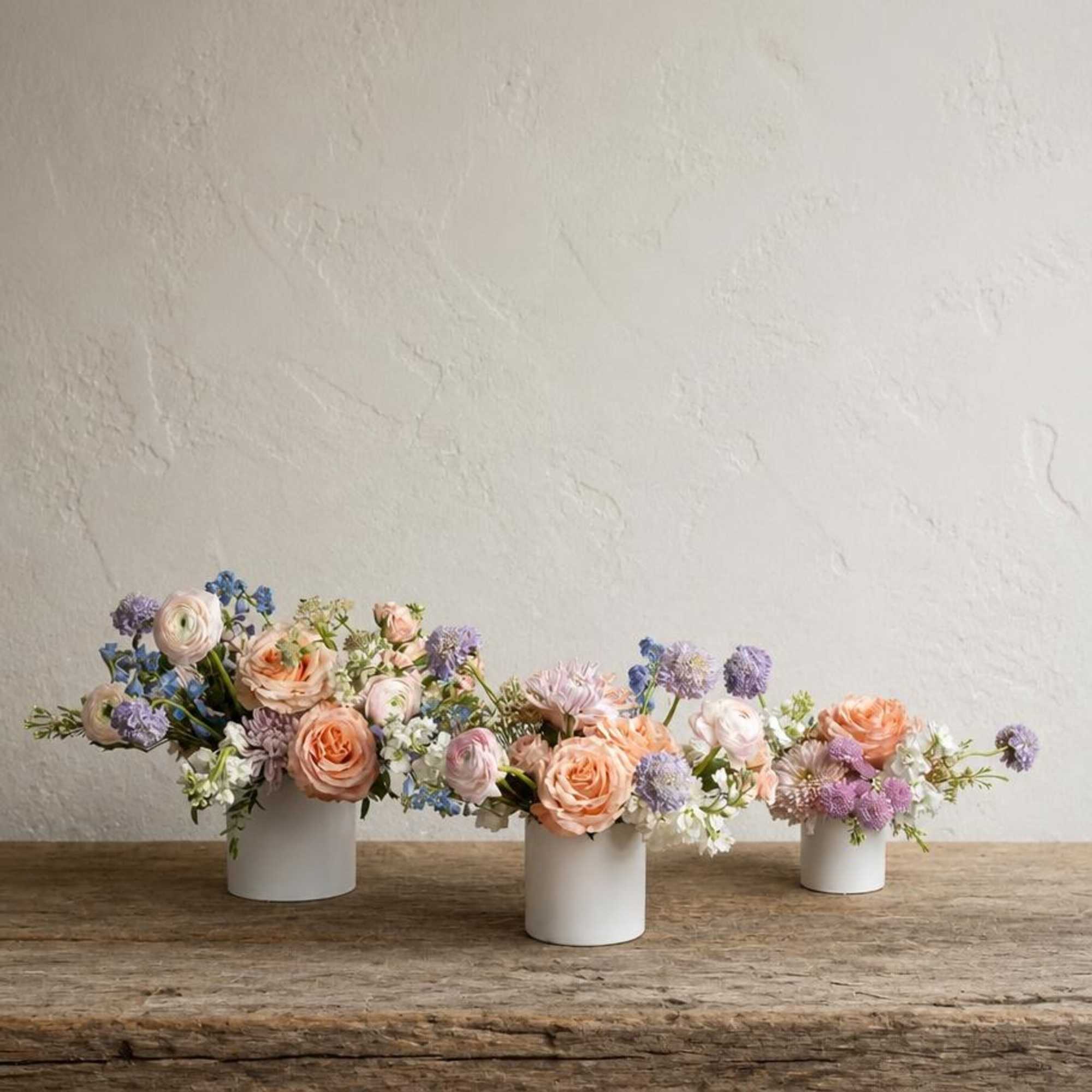 Three pastel floral arrangements in white cylindrical vases on a wooden table