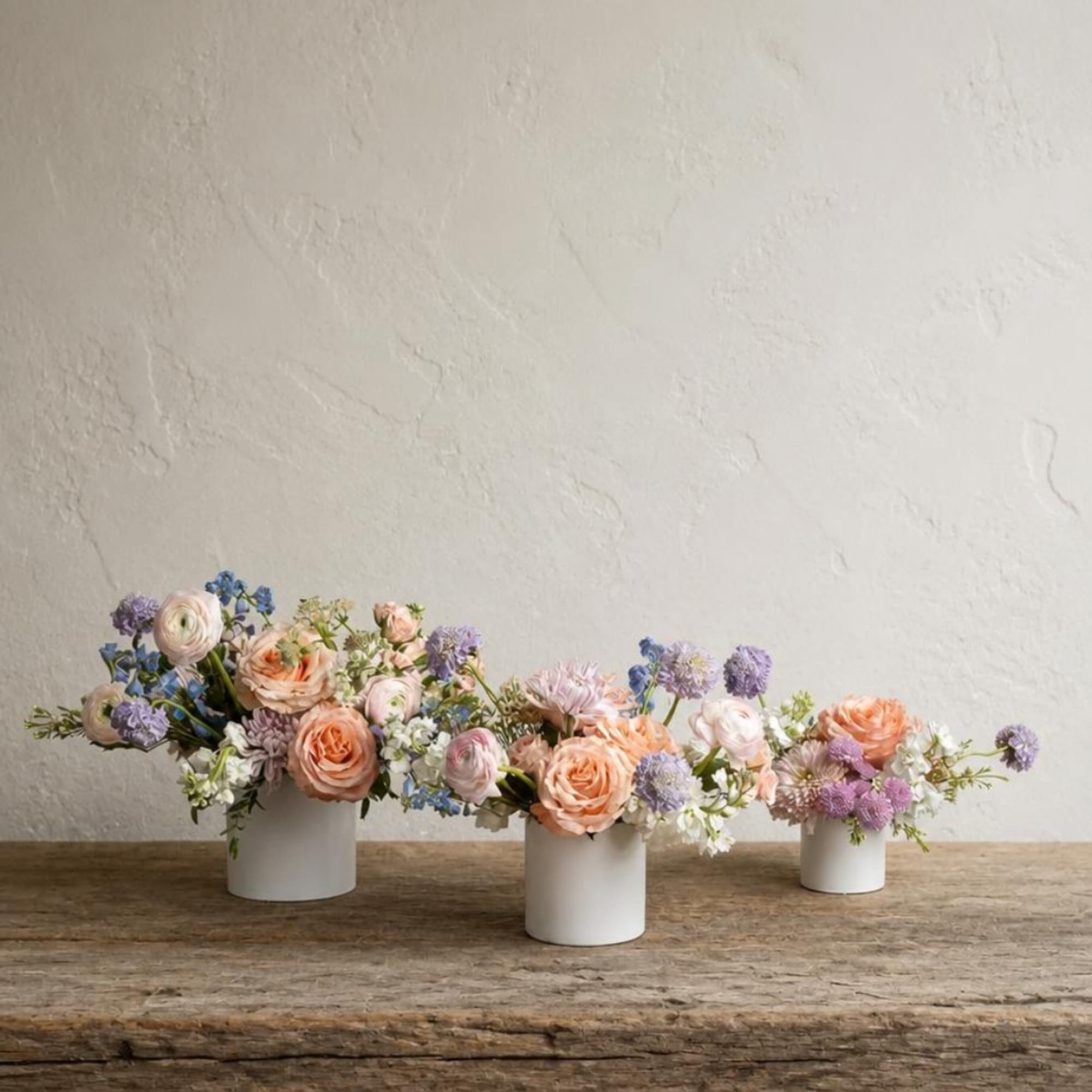Three pastel floral arrangements in white cylindrical vases on a wooden table