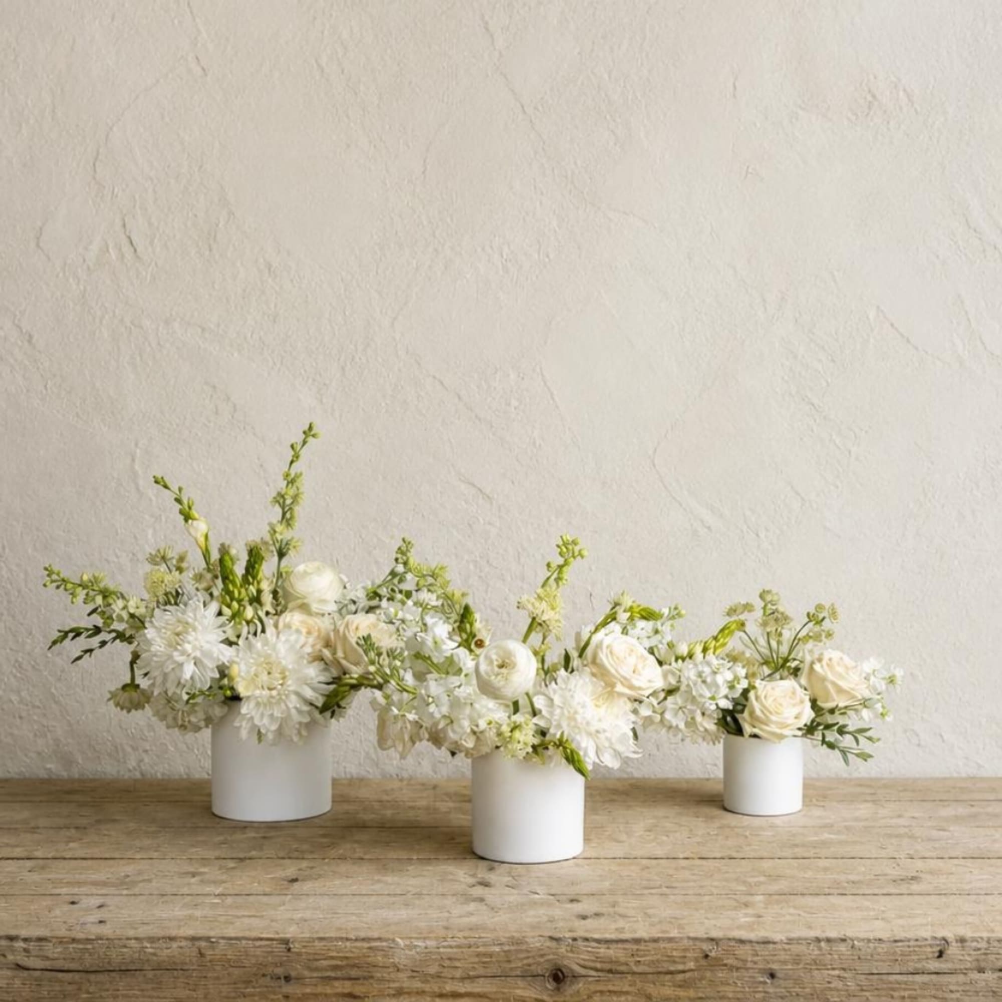 Three white floral arrangements in small white vases on a wooden table