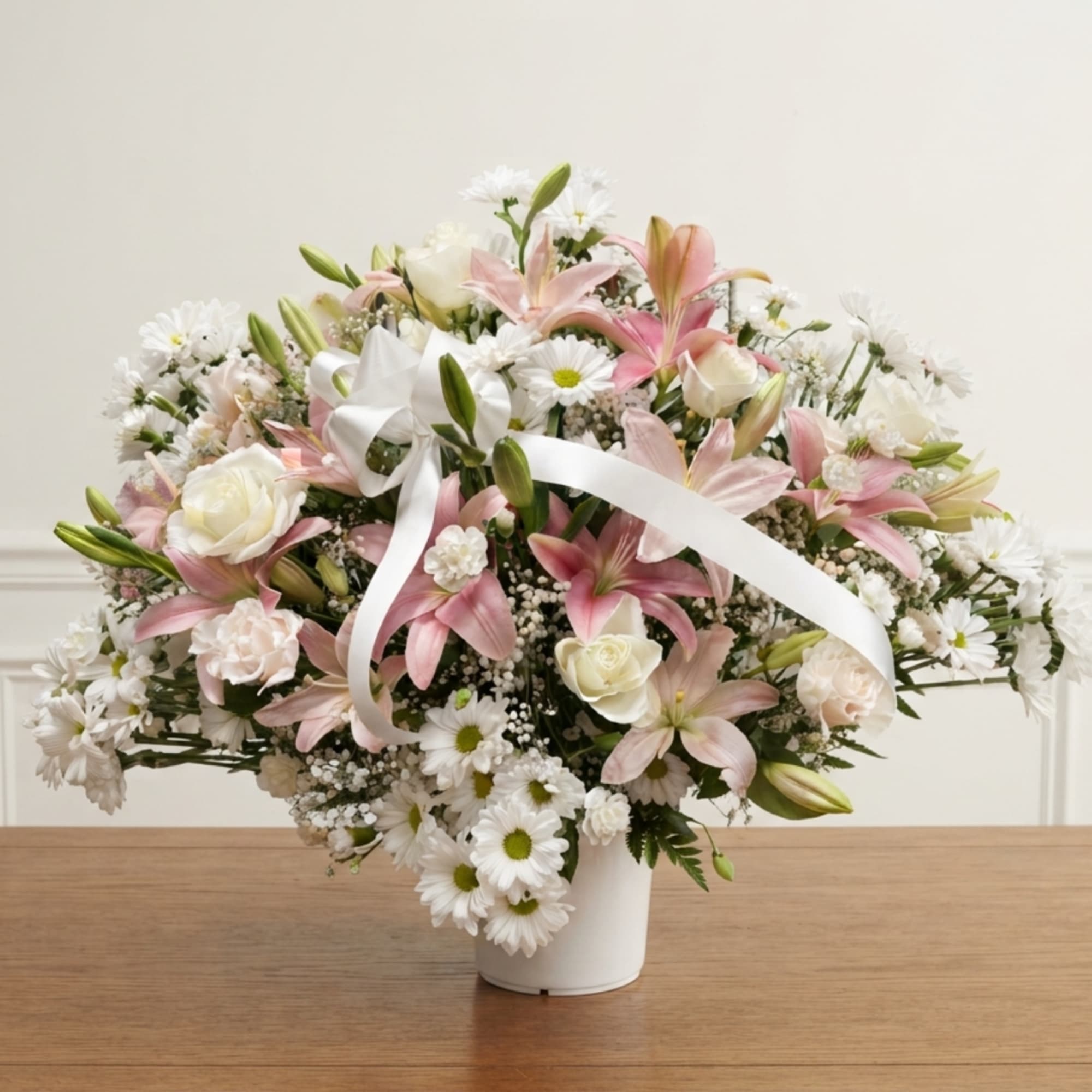 Pink lilies and white daisies arranged in a white vase with a ribbon