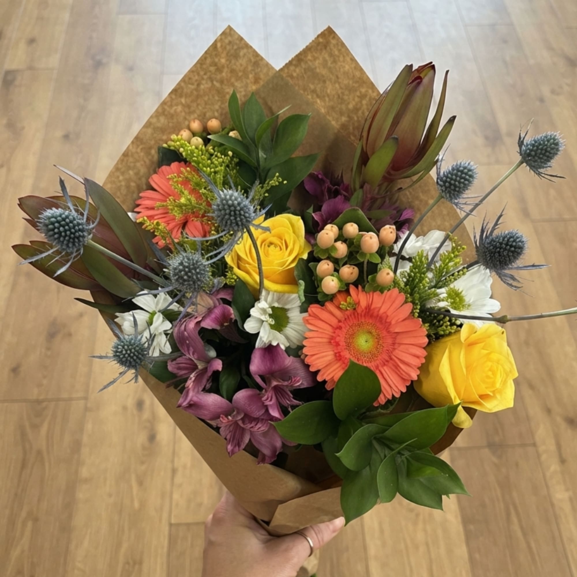 Handheld bouquet with yellow roses, orange gerberas, and purple blooms in brown paper