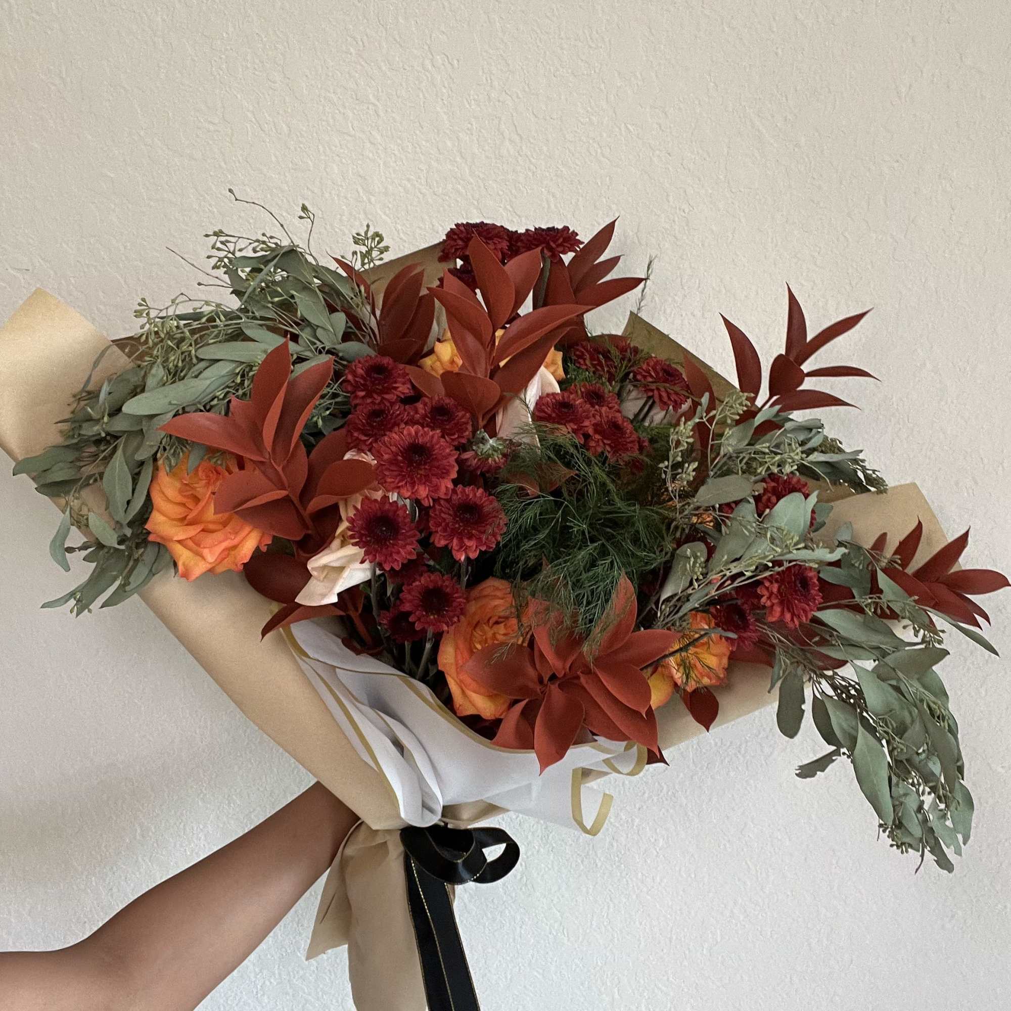 Handheld bouquet of orange roses and burgundy flowers wrapped in kraft paper
