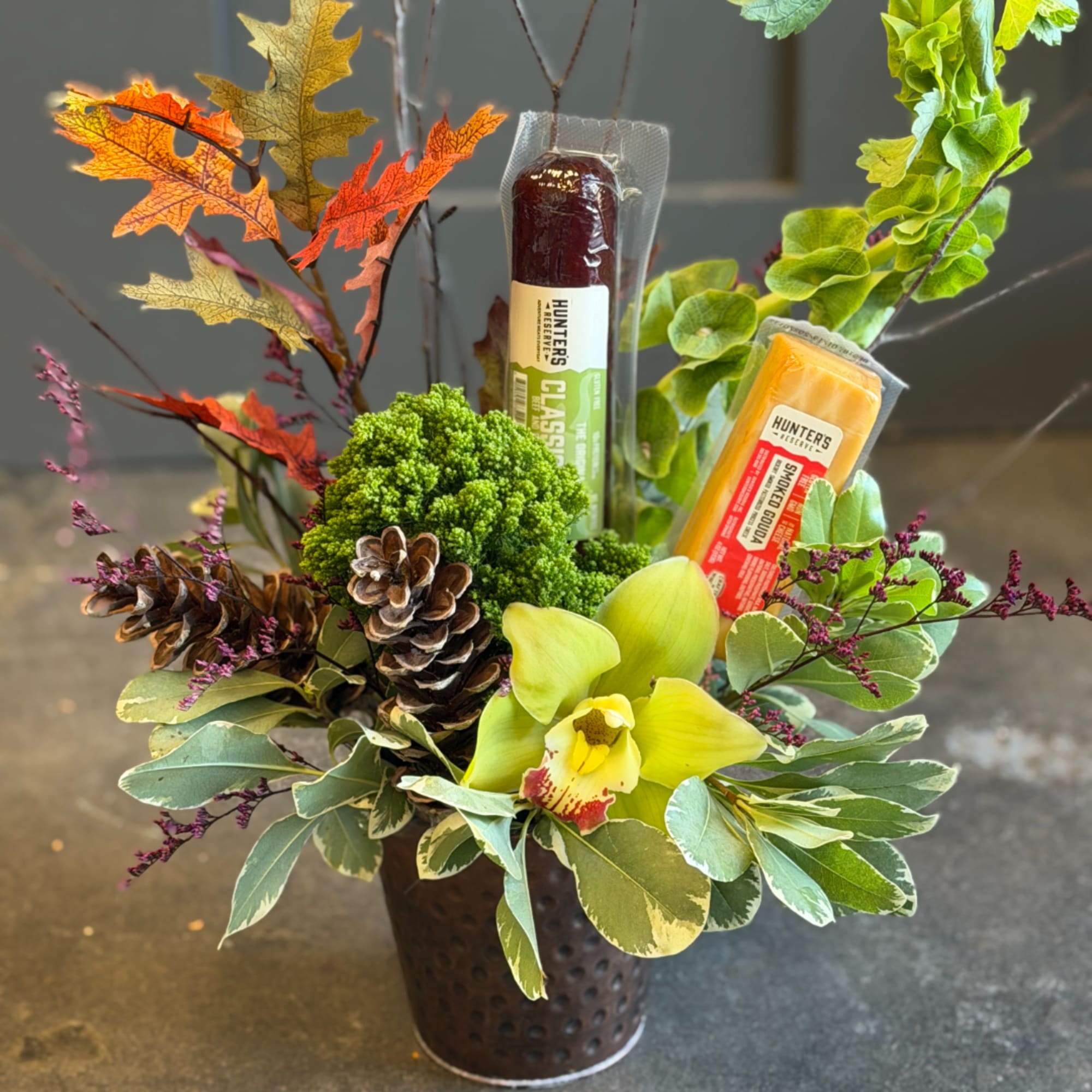 Autumn arrangement in a metal container with an orchid and packaged cheese sticks