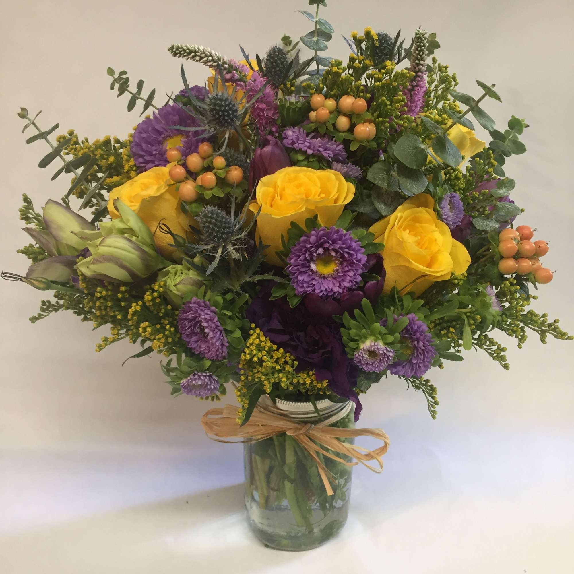 Bright yellow roses and purple asters arranged with berries in a mason jar tied with raffia