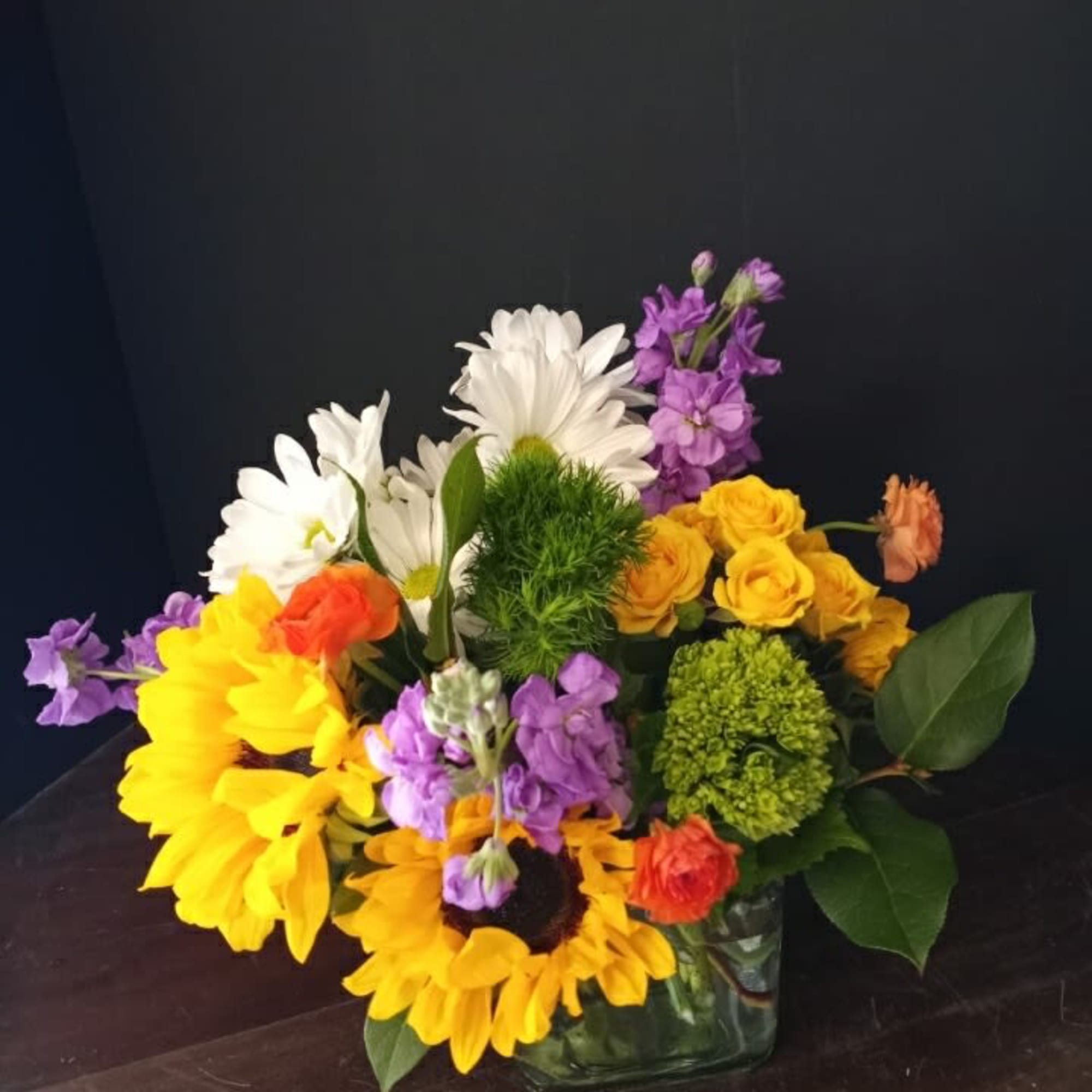 Mixed bouquet of sunflowers, white daisies, and purple flowers in a glass vase
