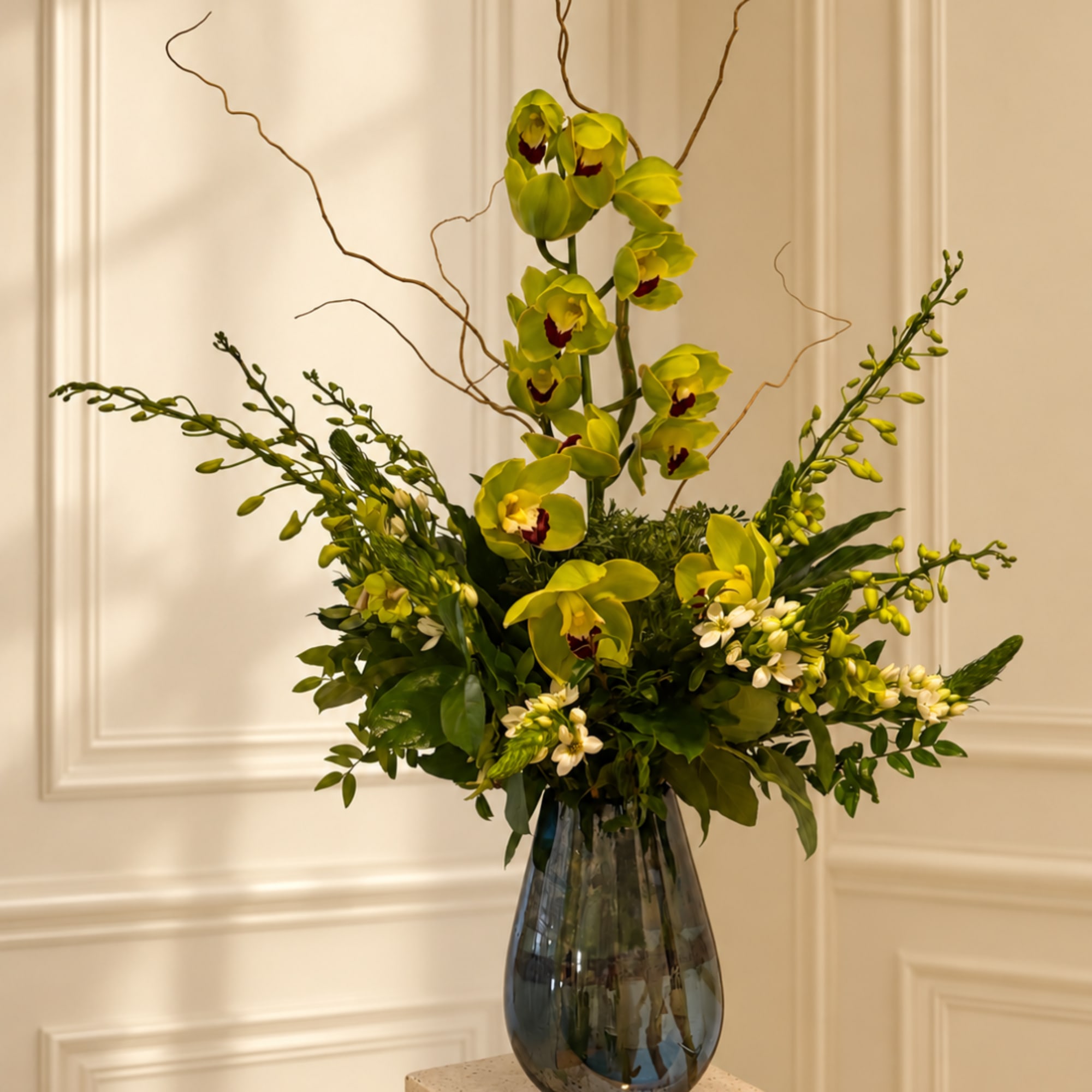 Tall floral arrangement with green orchids and white accent flowers in a glass vase