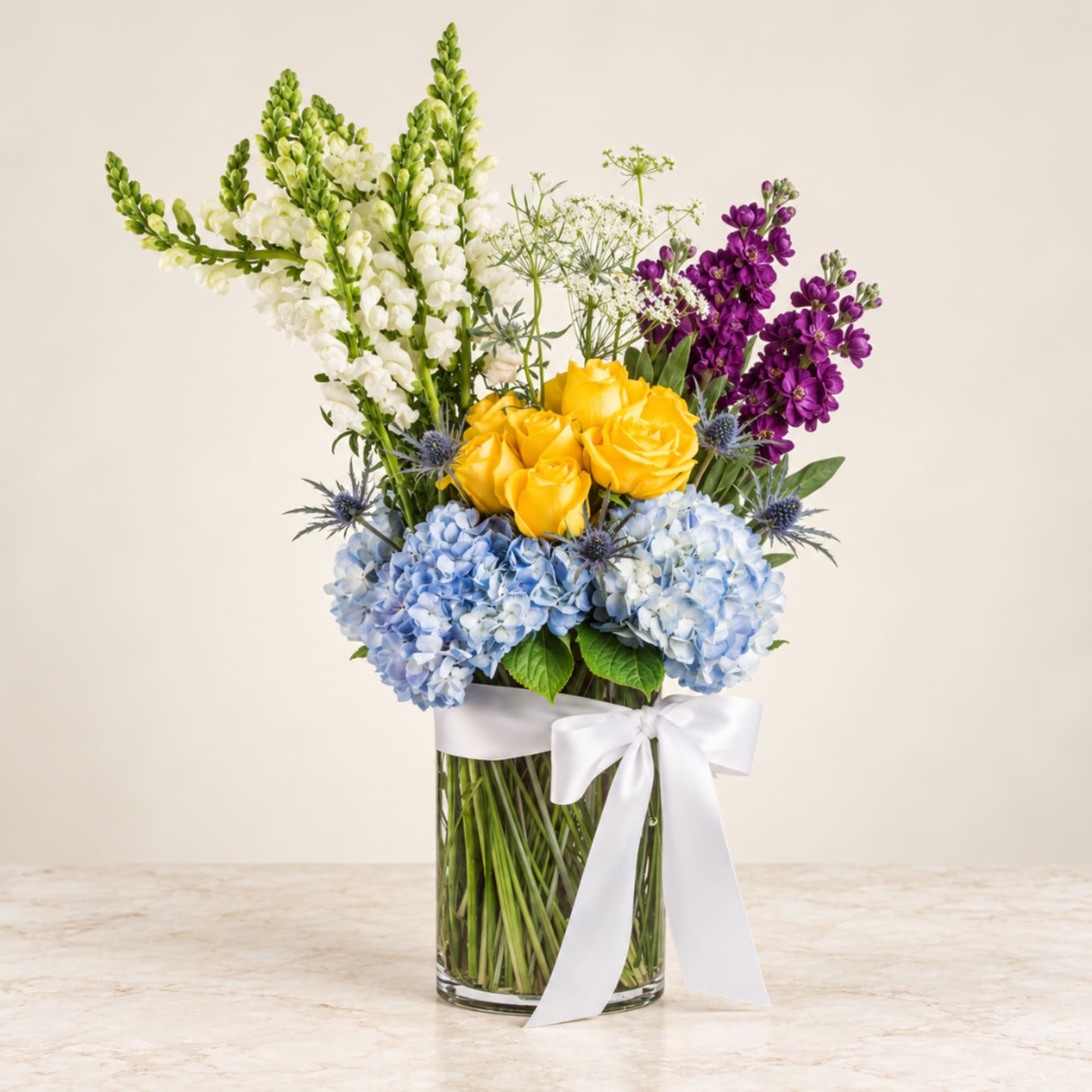 Mixed bouquet in a glass vase with yellow roses, blue hydrangeas, and purple flowers