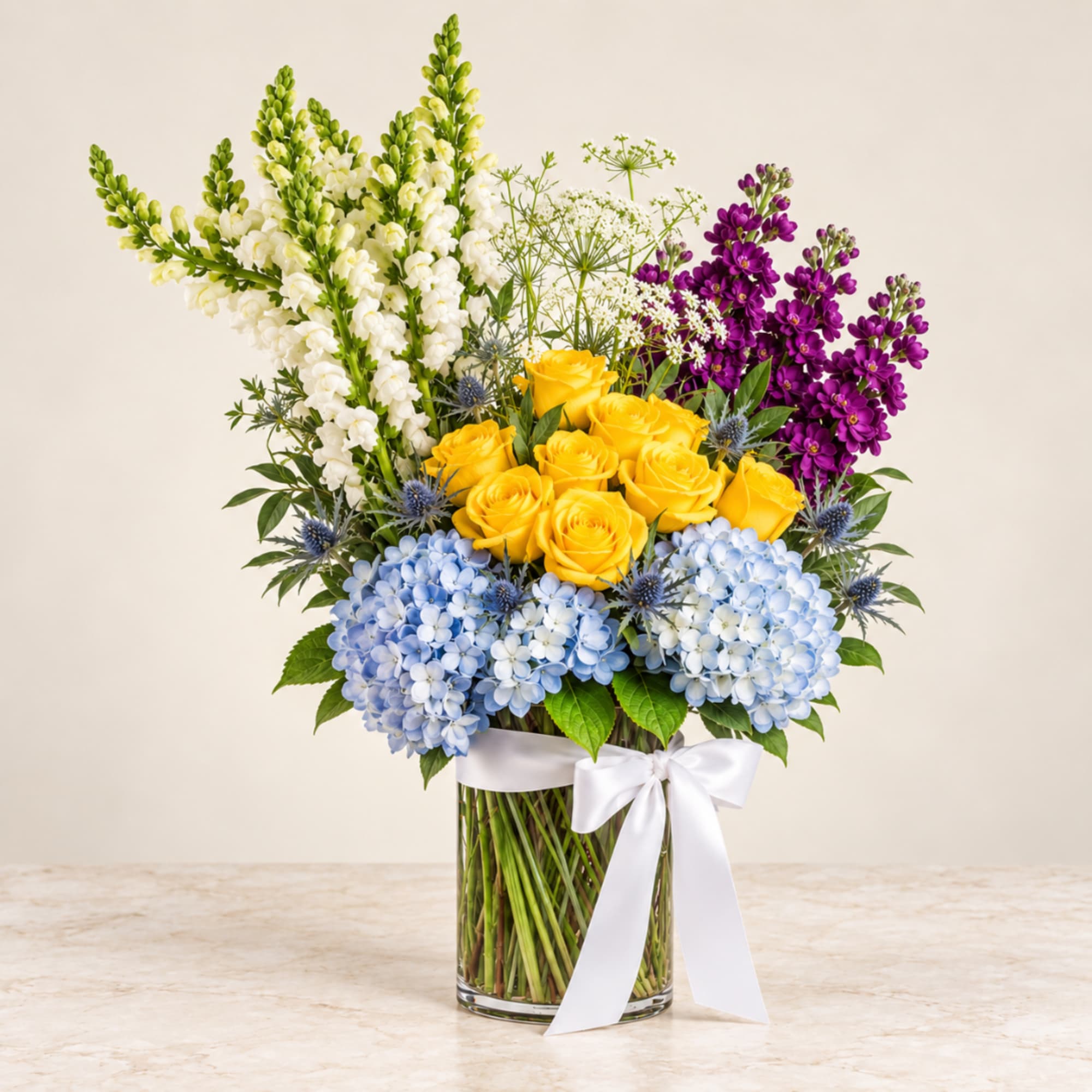 Bouquet of yellow roses, blue hydrangeas, white snapdragons, and purple flowers in a glass vase