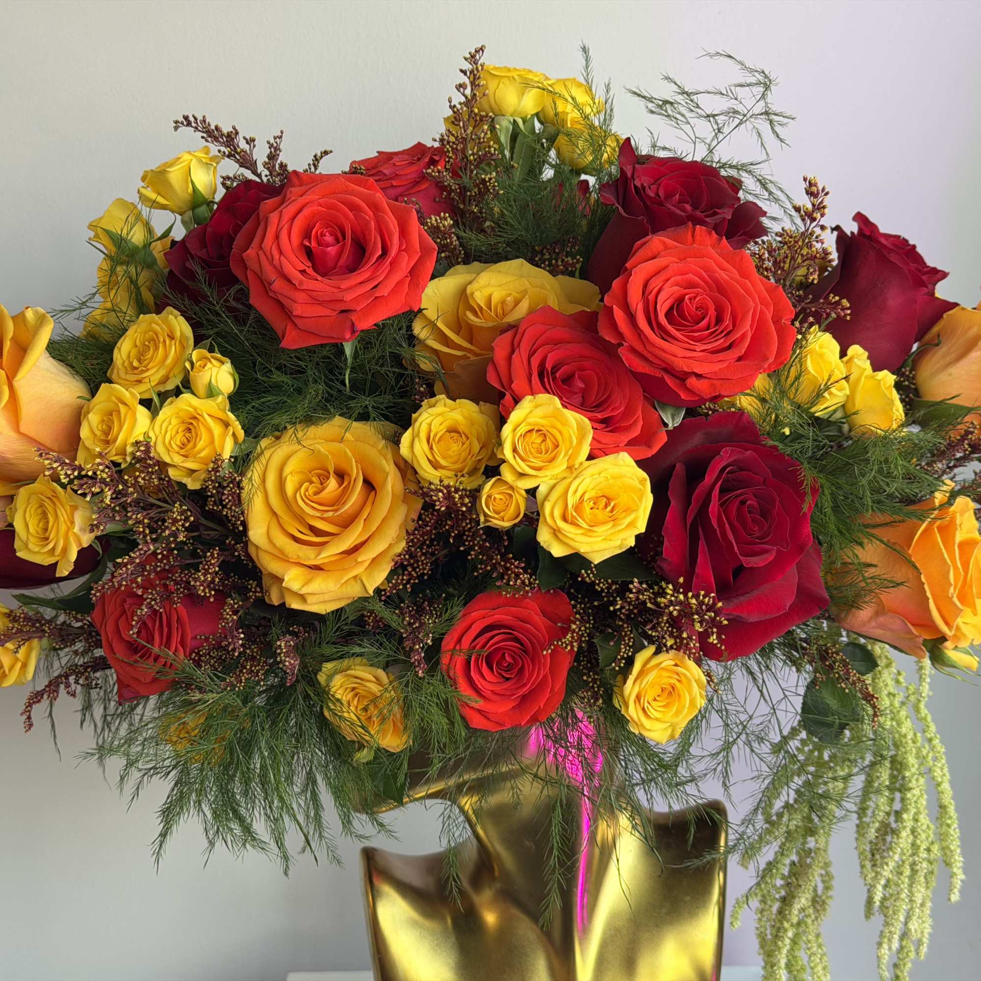 Arrangement of red, yellow, and orange roses in a gold vase with trailing greenery