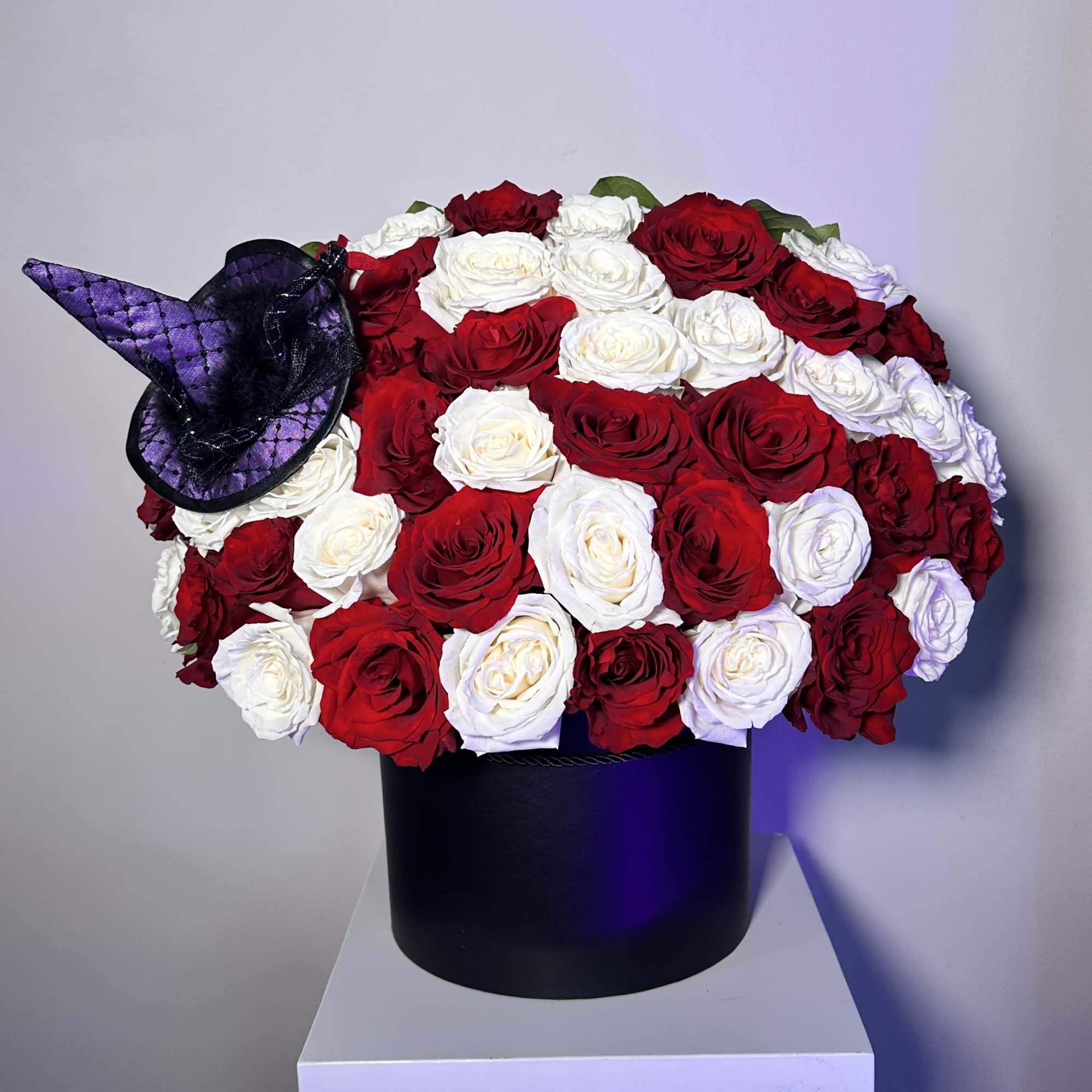 Round arrangement of red and white roses in a black hat box with a small purple witch hat accent.