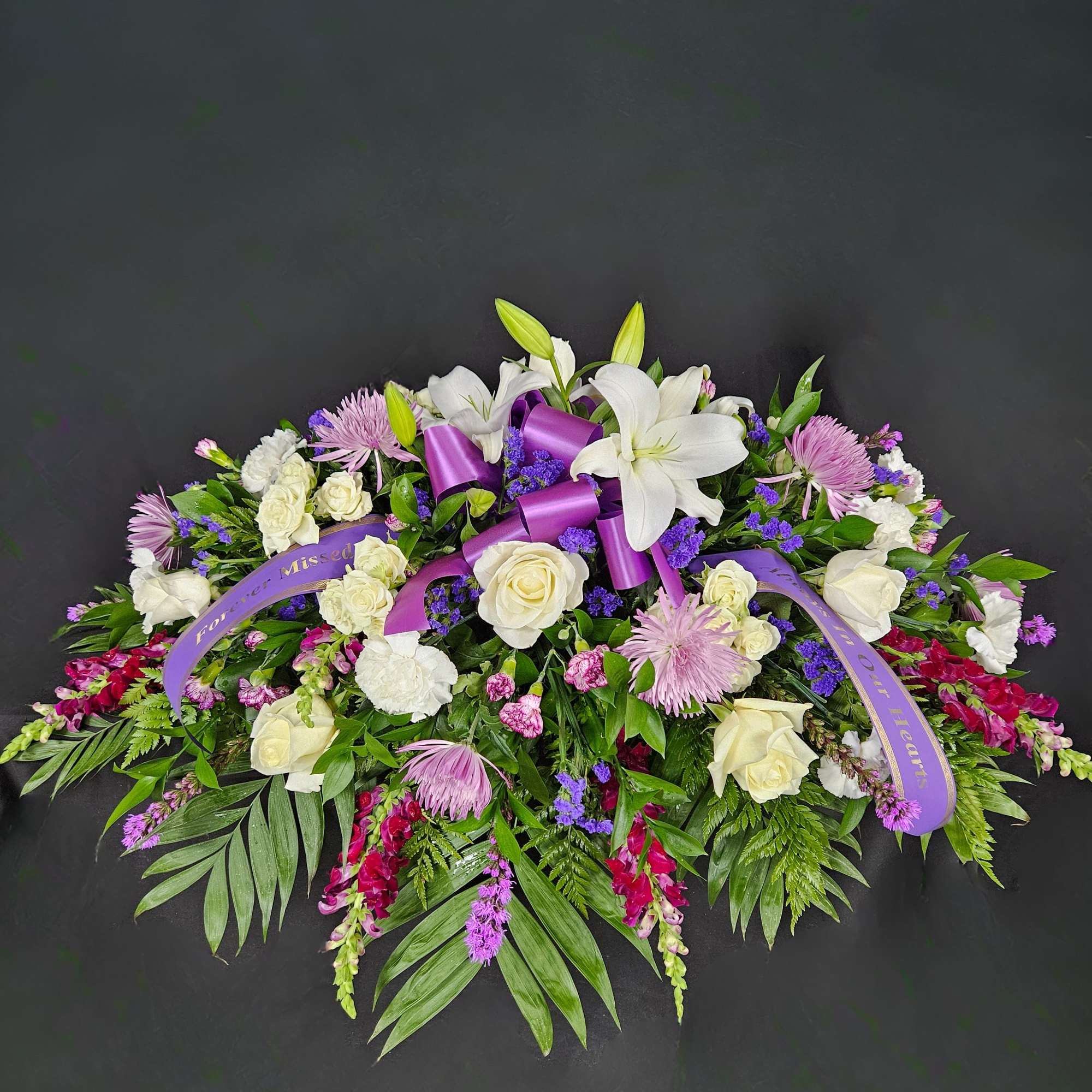 Elongated spray of white lilies, roses and purple flowers with a purple sympathy ribbon on a dark background