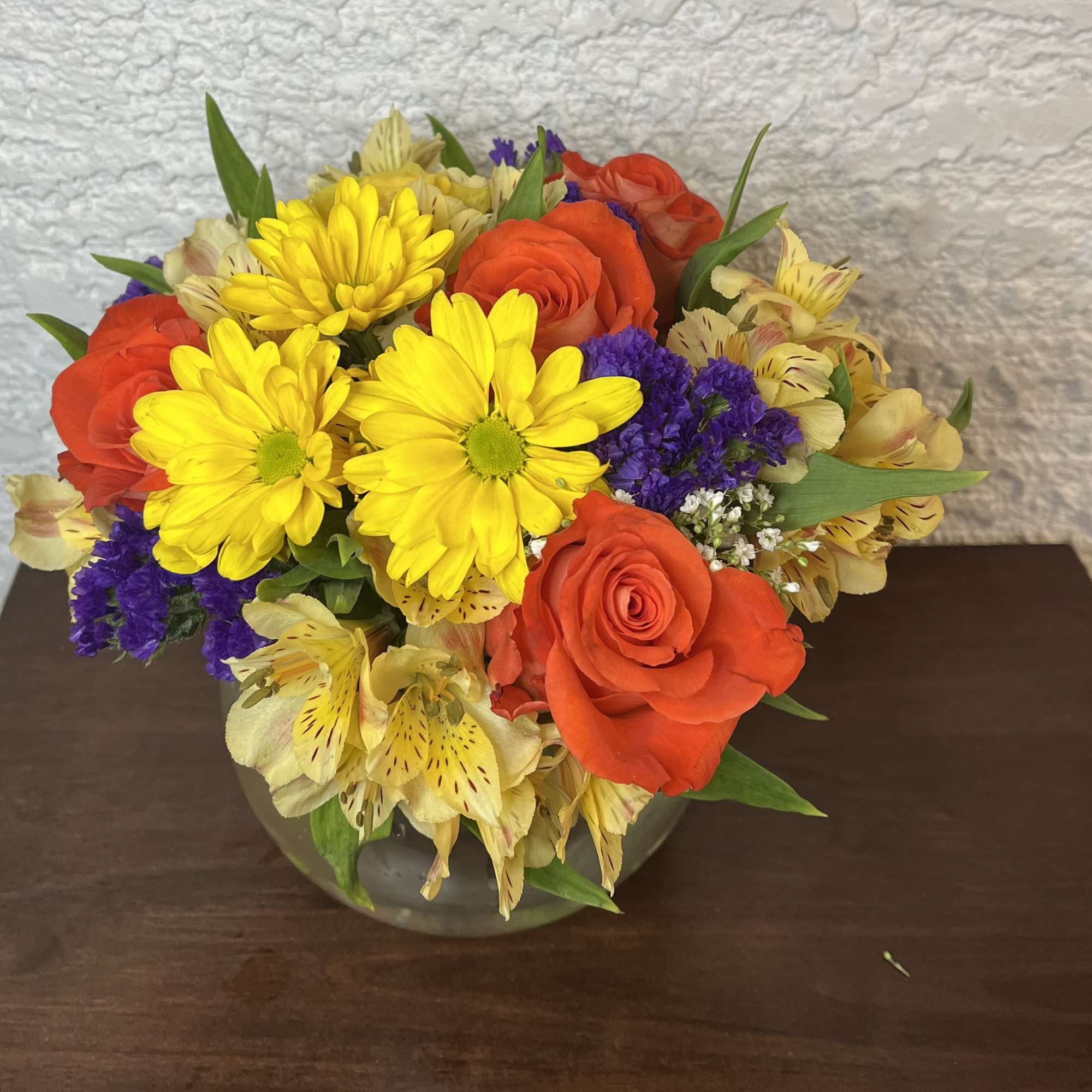 Bouquet of yellow daisies, orange roses, and purple filler in a glass vase