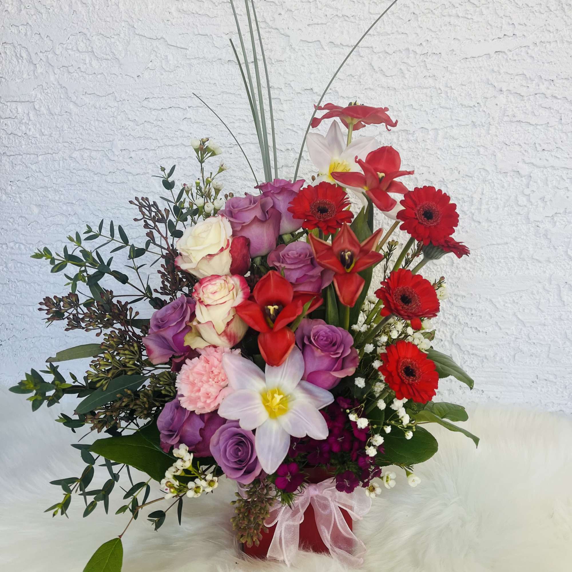 Tall bouquet of red, purple, and white flowers in a red vase with a pink ribbon