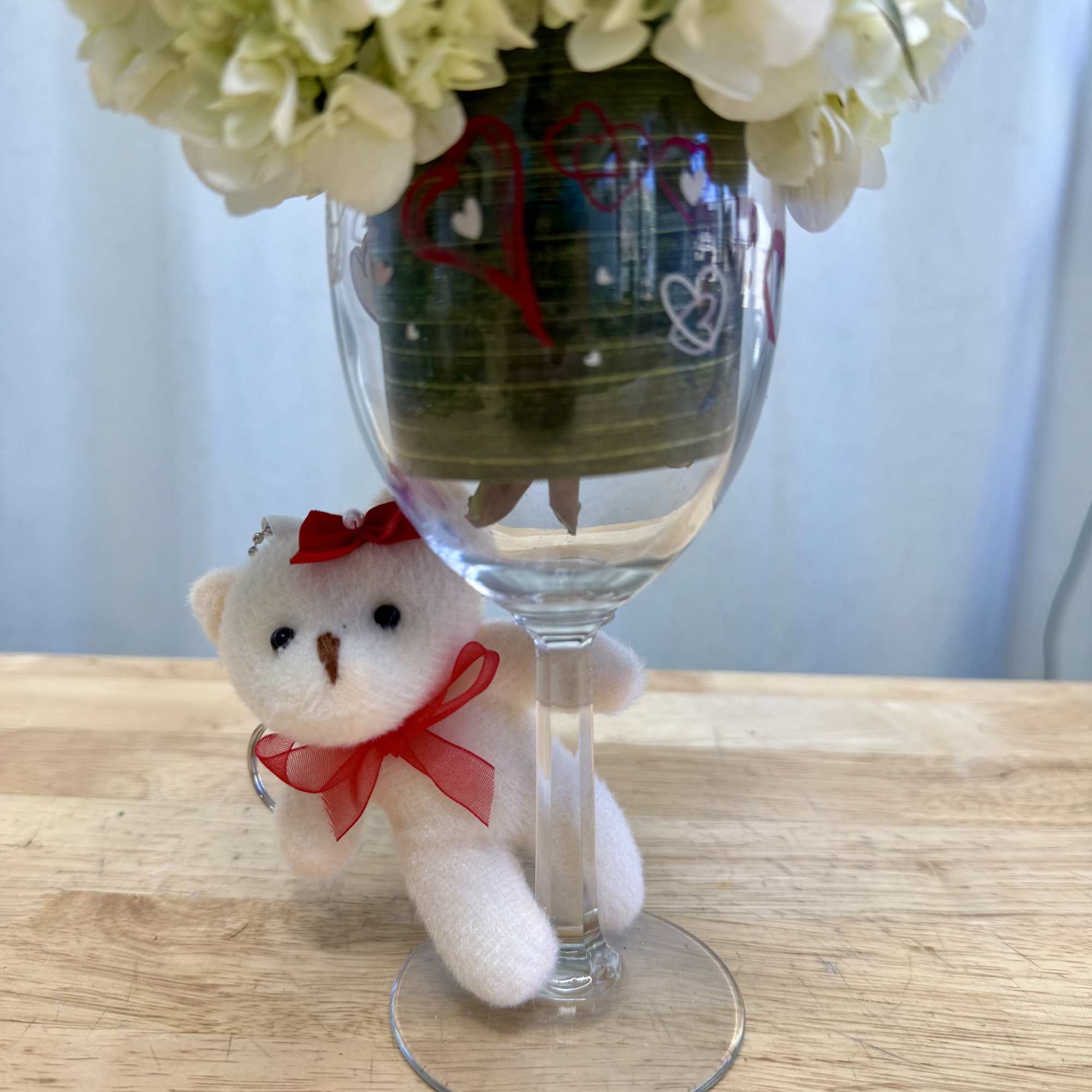 White hydrangea arrangement in a wine glass with a small teddy bear charm