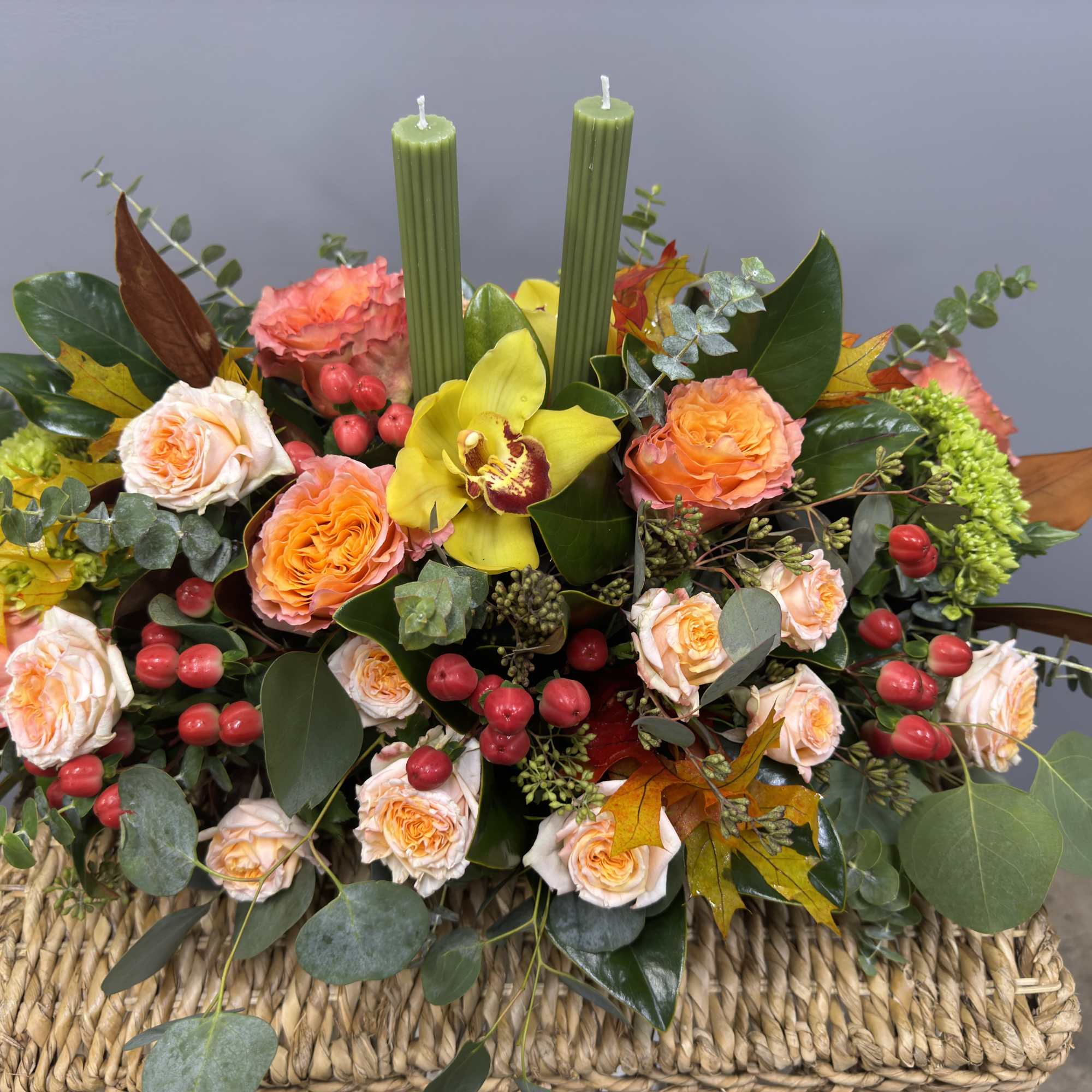 Autumn floral arrangement with roses, a yellow orchid, and two green candles in a woven basket