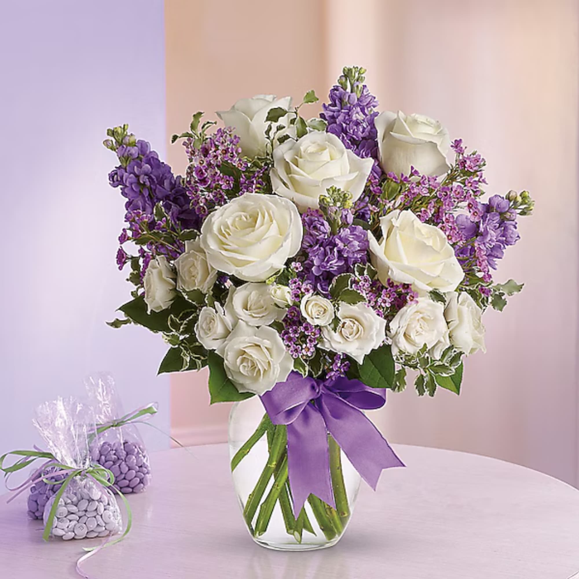 White roses and purple flowers arranged in a clear glass vase with a purple ribbon.