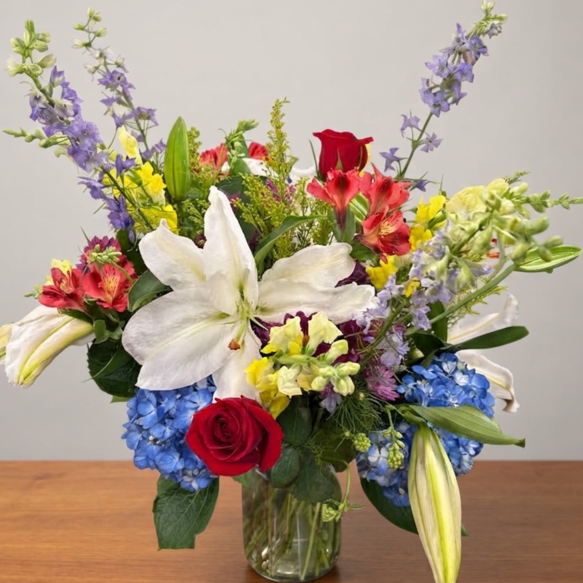 Mixed bouquet with white lilies, red roses, blue hydrangeas, and purple flowers in a glass vase