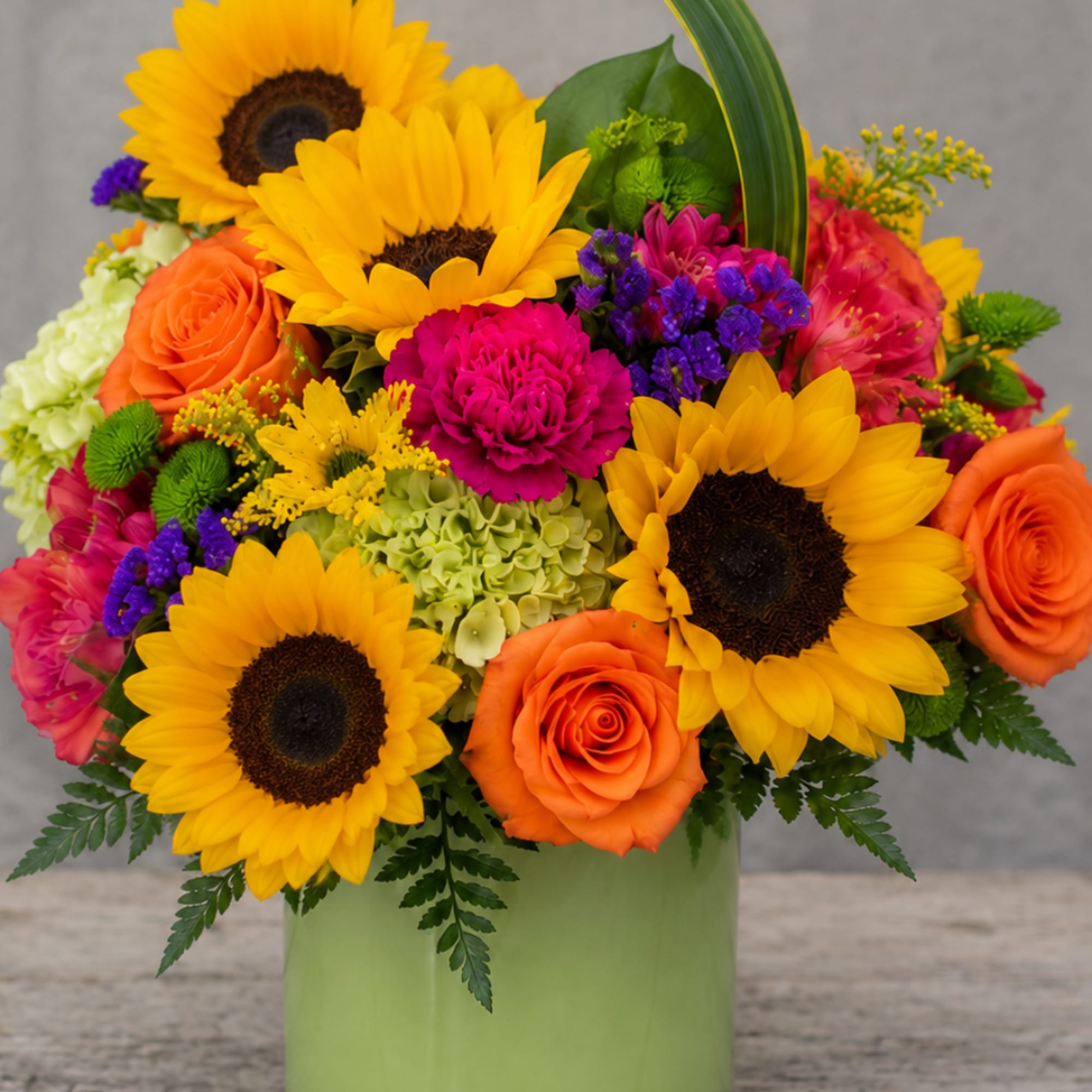 Bright bouquet of sunflowers, orange roses, and pink blooms in a green vase