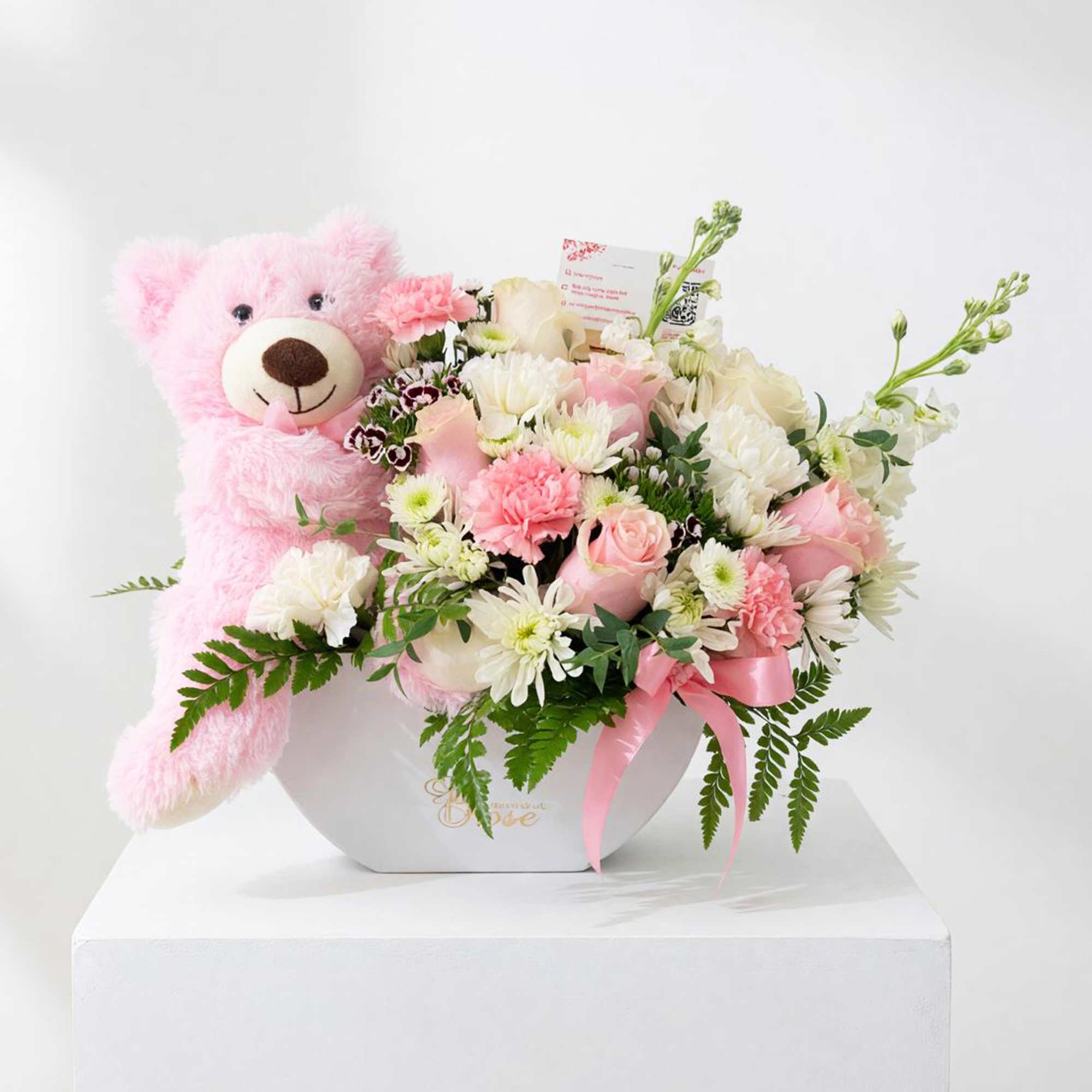 Pink teddy bear beside a pastel flower arrangement in a white box
