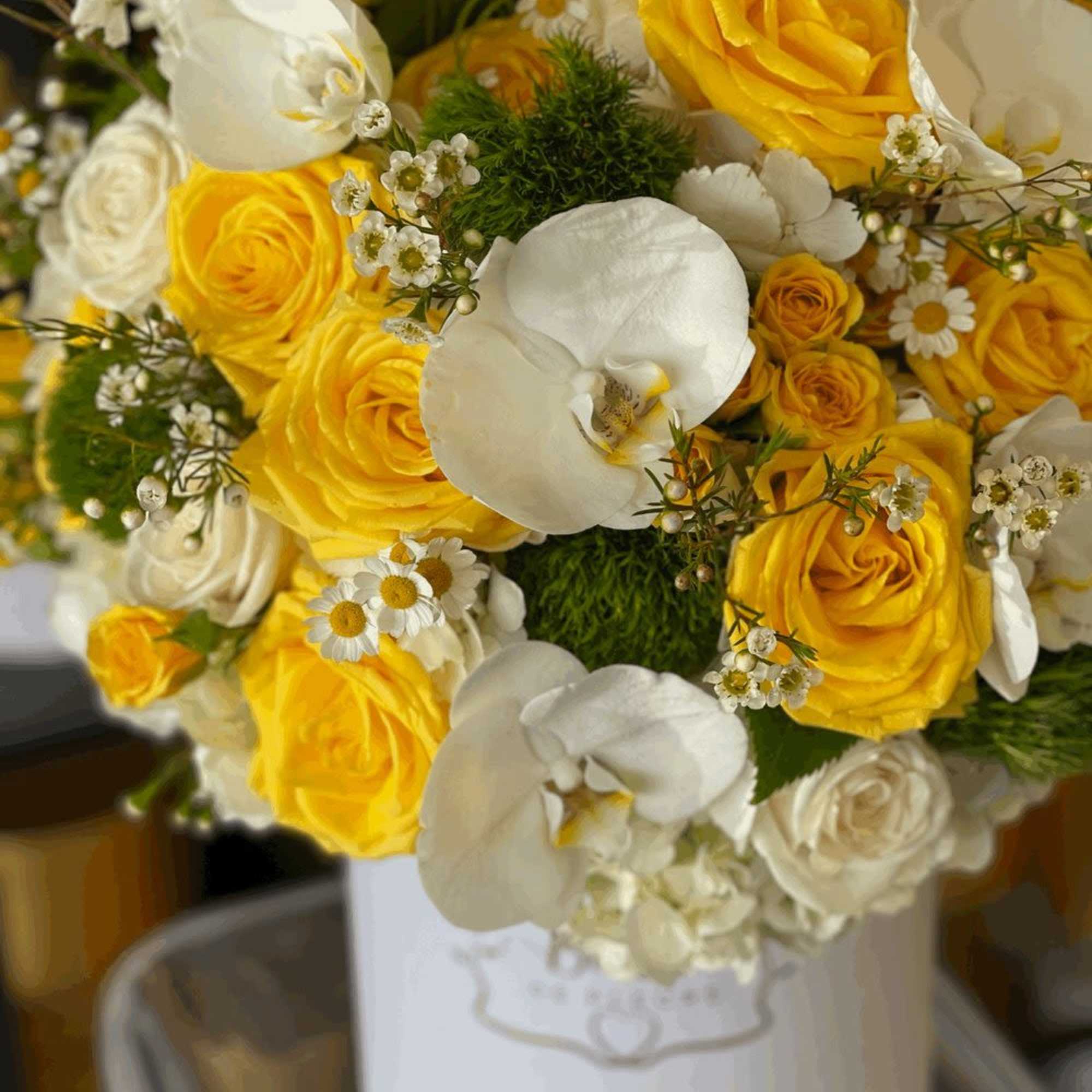 Yellow and white roses with white orchids and daisies in a round white hatbox.