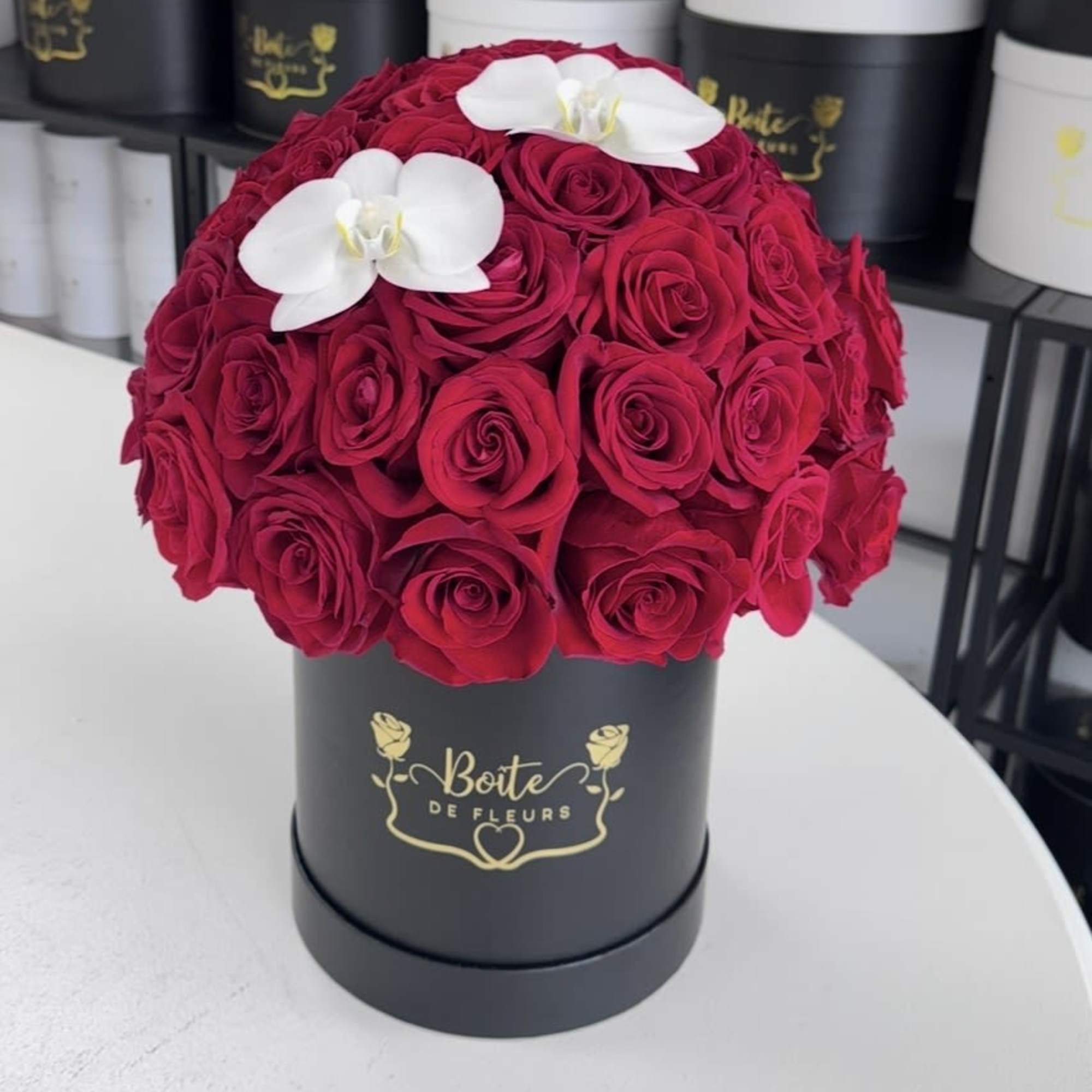Red roses arranged in a black round box with two white orchids