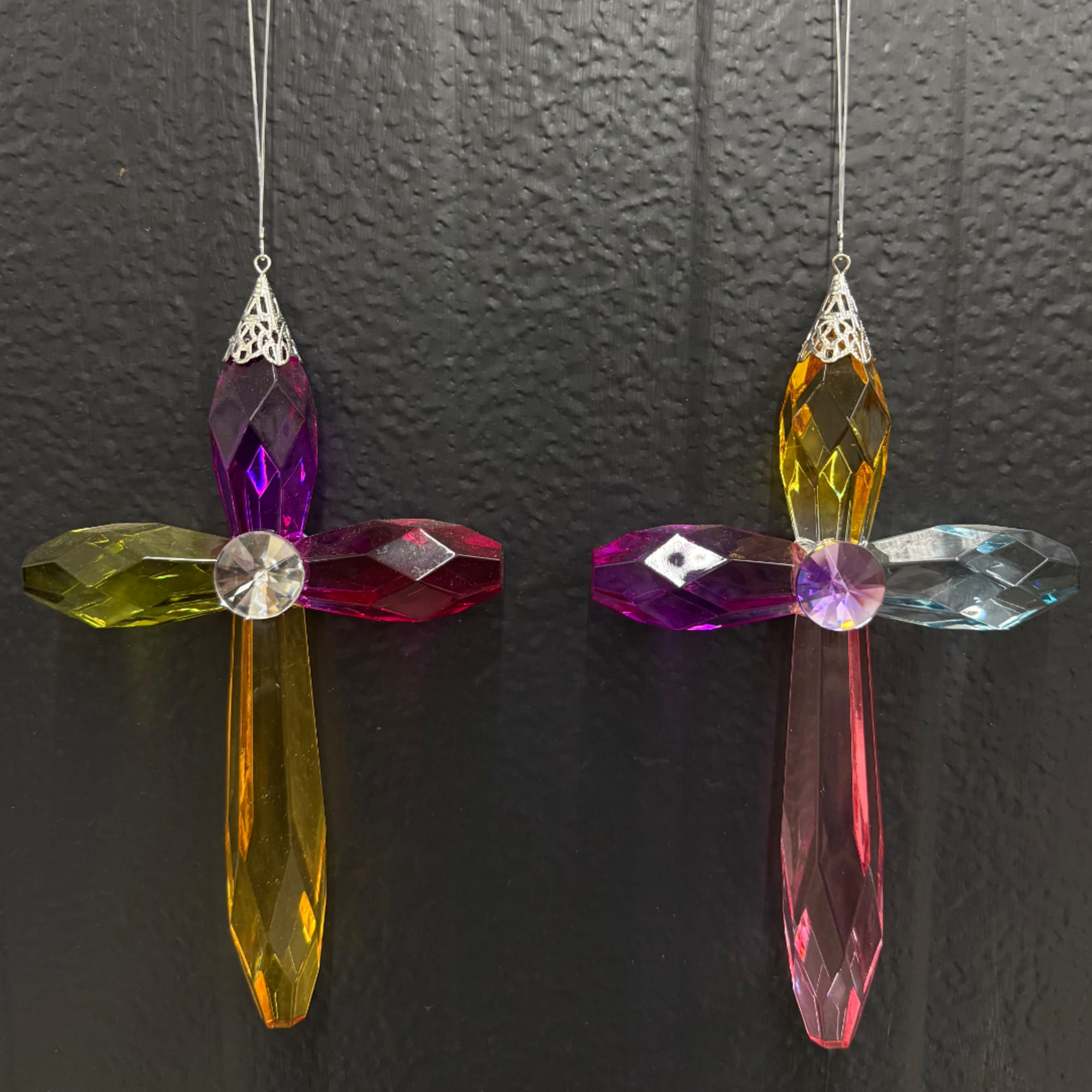 Two colorful crystal cross ornaments hang against a dark wall.