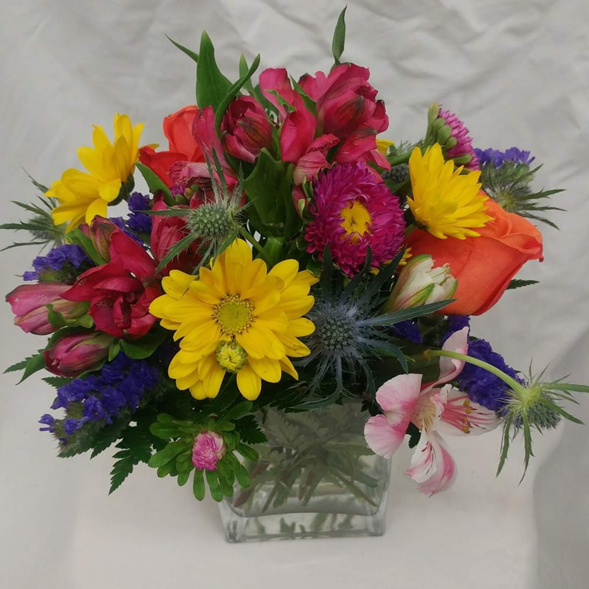 Mixed bouquet in a clear square vase with yellow daisies and pink, orange, and purple blooms