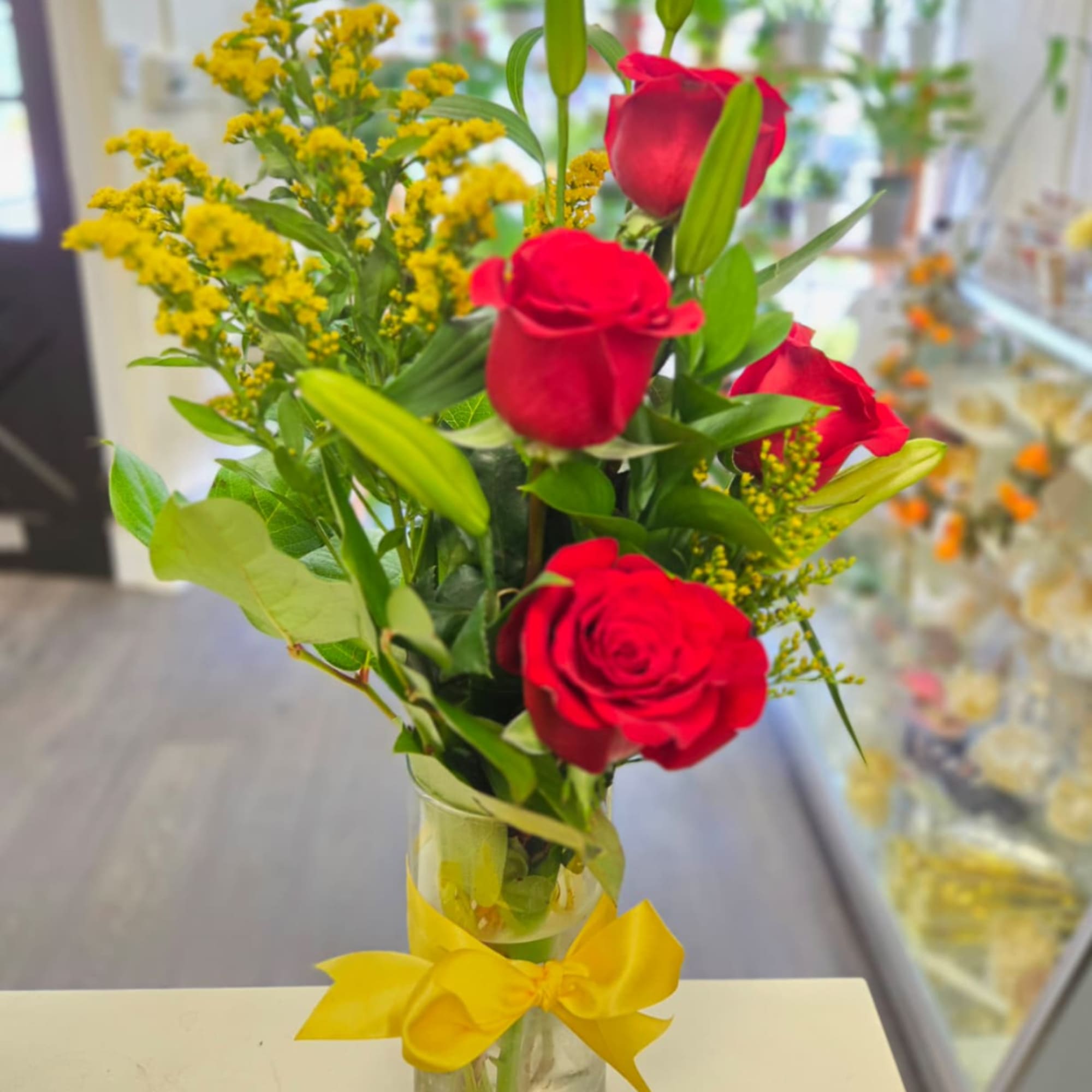 Red roses and yellow filler flowers in a clear vase with a yellow bow.