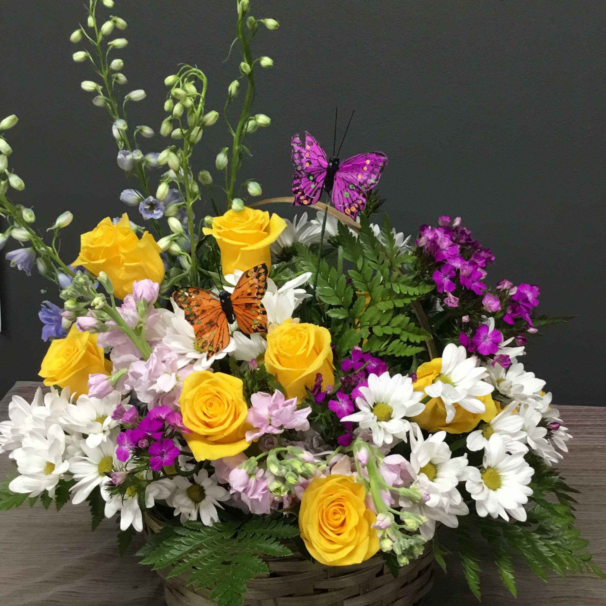 Basket arrangement of yellow roses, white daisies, and purple flowers with butterfly picks