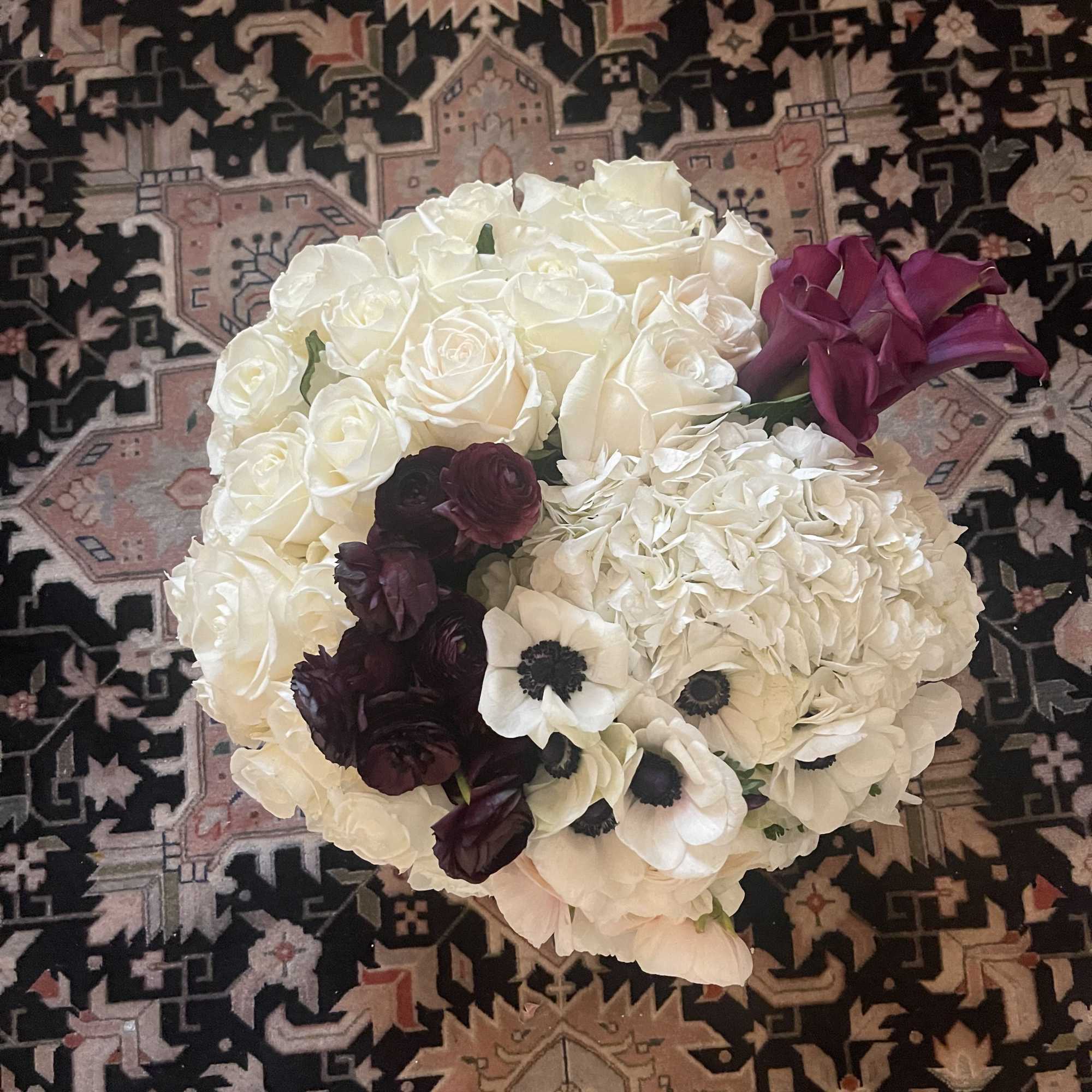 Round arrangement of white roses, hydrangeas, anemones, and dark burgundy and plum accent flowers.