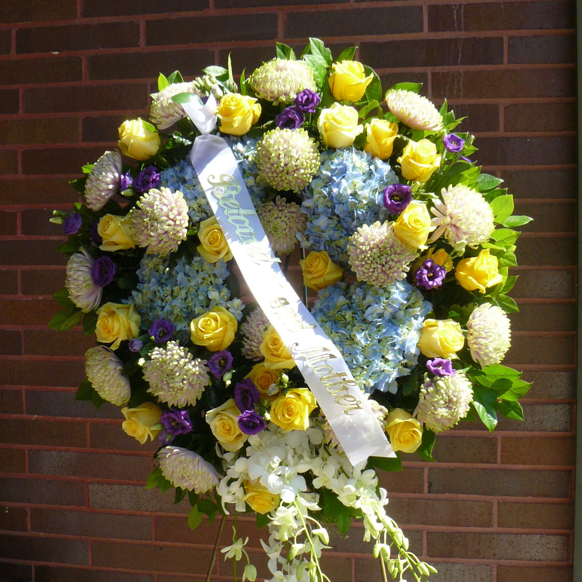 Large funeral wreath of yellow roses, blue hydrangeas, and purple flowers with a ribbon