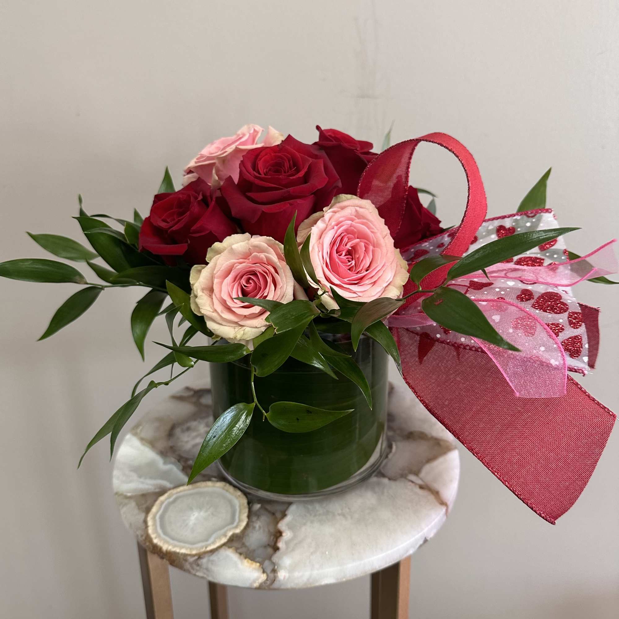 Short arrangement of red and pink roses with greenery in a glass vase, accented with a red and pink bow.