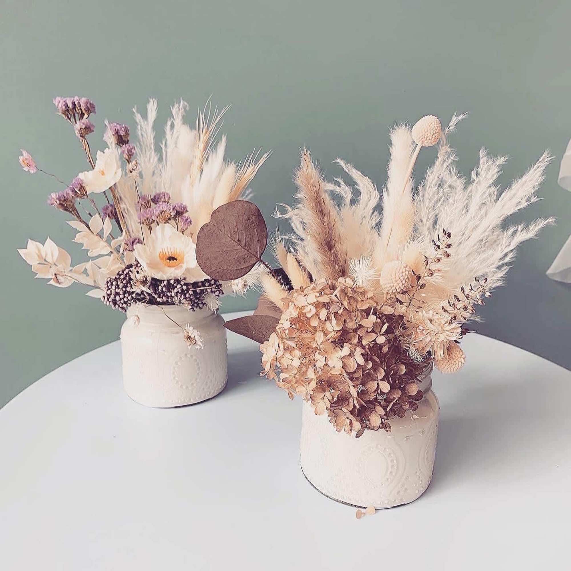 Two dried floral arrangements in white ceramic jars on a table