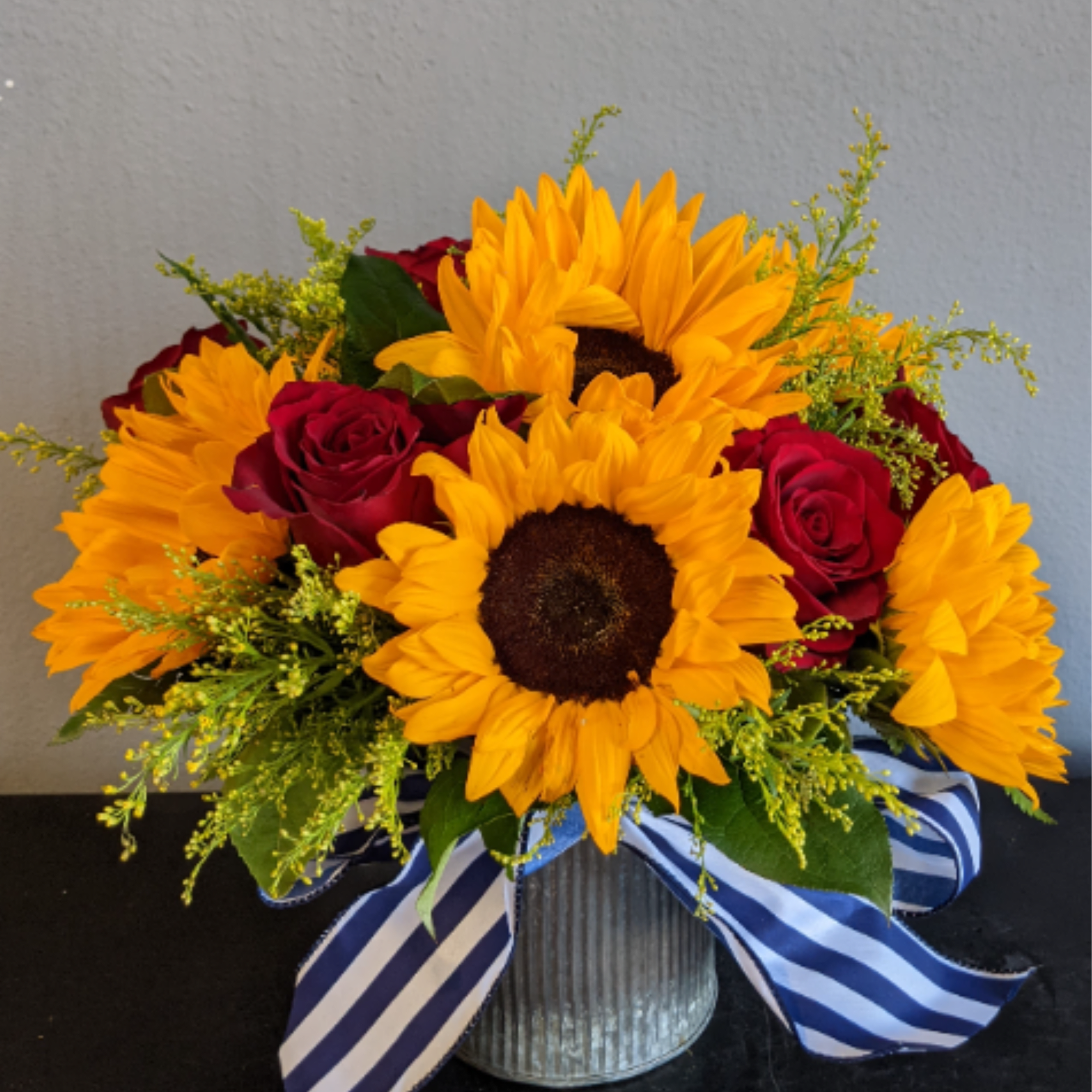 Low arrangement of bright sunflowers and red roses in a metal vase with a striped blue ribbon.