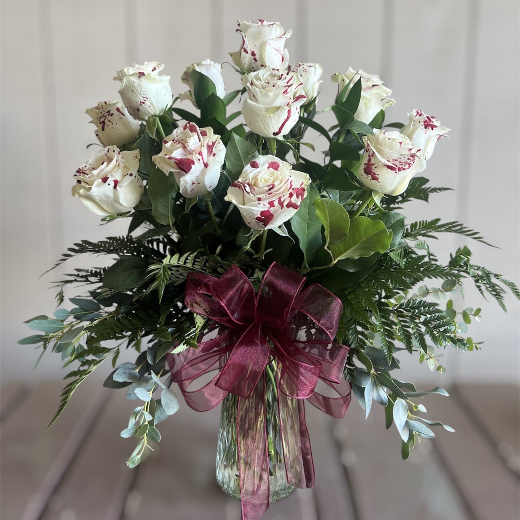 White roses with red speckles in a glass vase with a burgundy ribbon