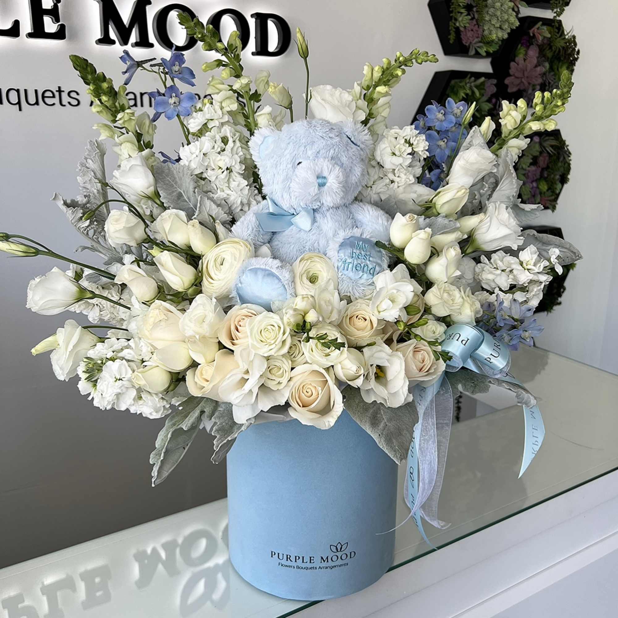 Blue teddy bear in a white and blue flower arrangement in a round box