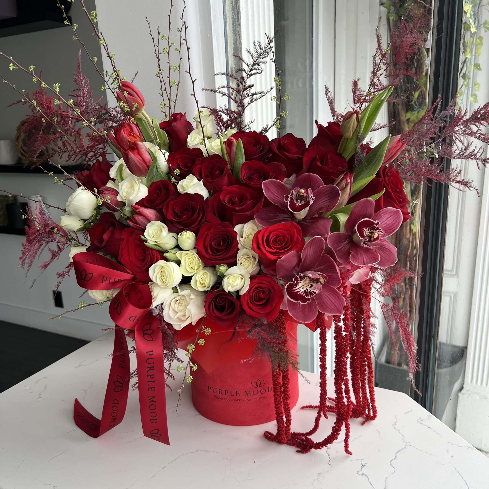 Large red and white rose arrangement with mauve orchids in a red hatbox with cascading red accents.