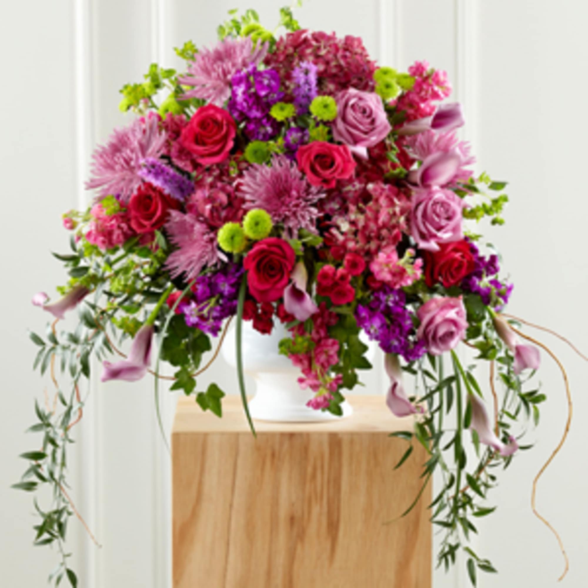 Lush pink, purple, and red flower arrangement with cascading greenery in a white pedestal vase.