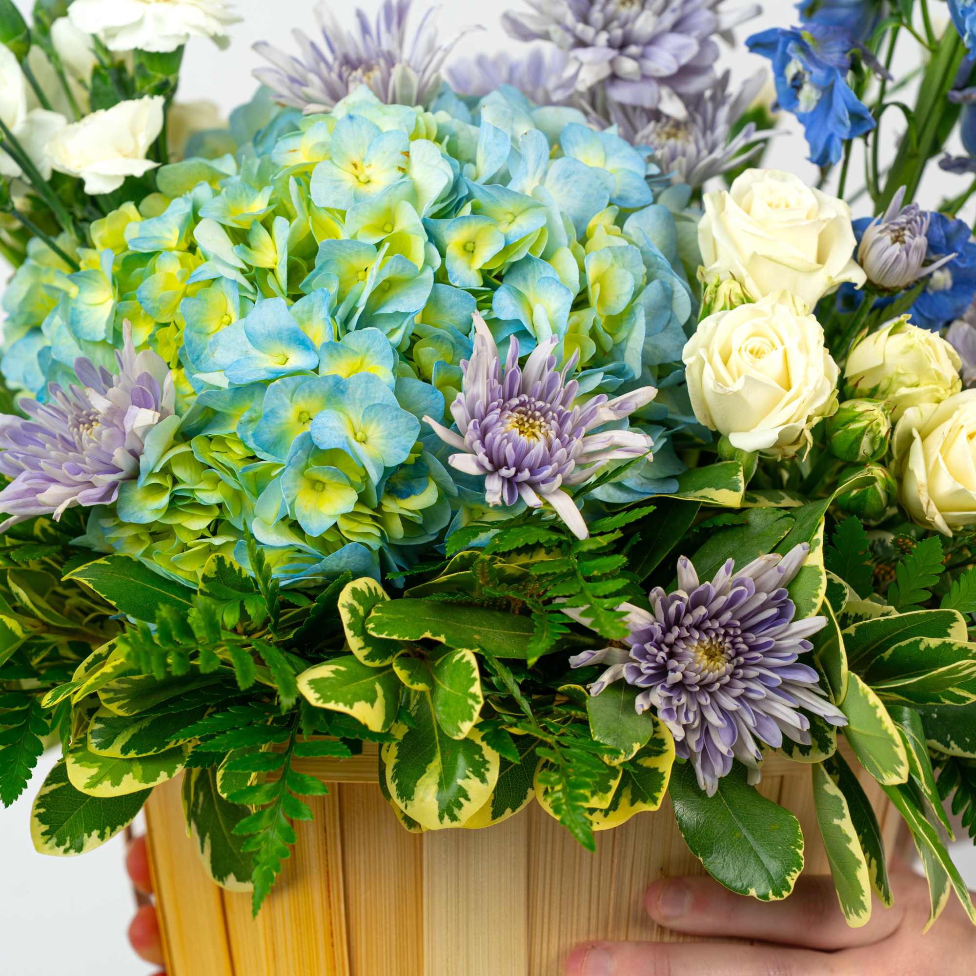 Arrangement of blue hydrangeas, white roses, purple mums, and blue delphinium in a wooden container.