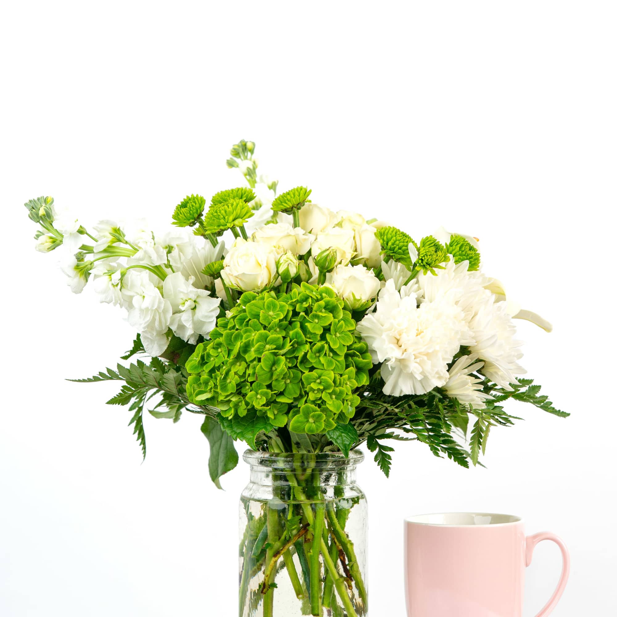 White and green flower arrangement in a glass jar beside a pink mug on a white surface.