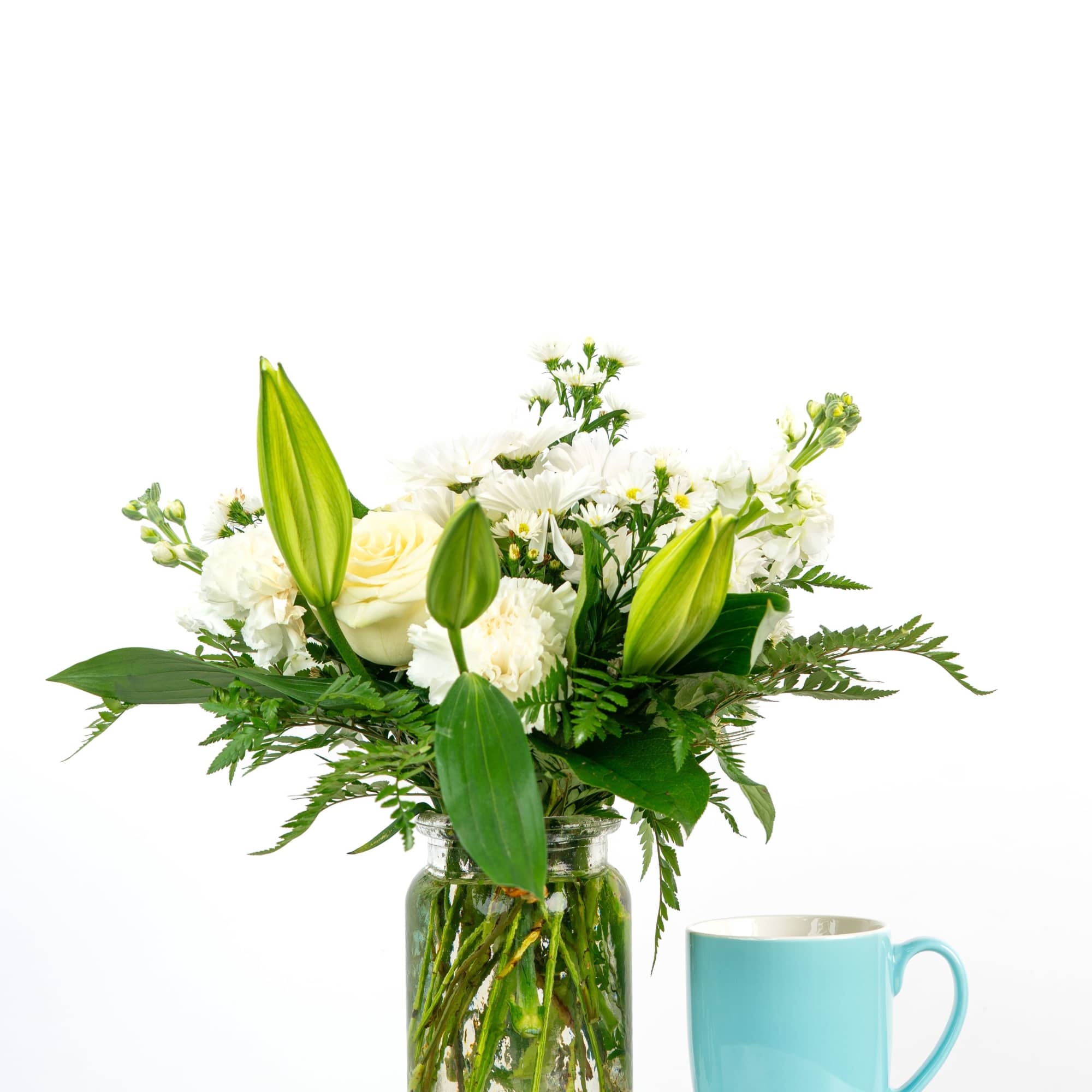 White lilies, roses, carnations, and mums in a clear glass jar beside a light blue mug